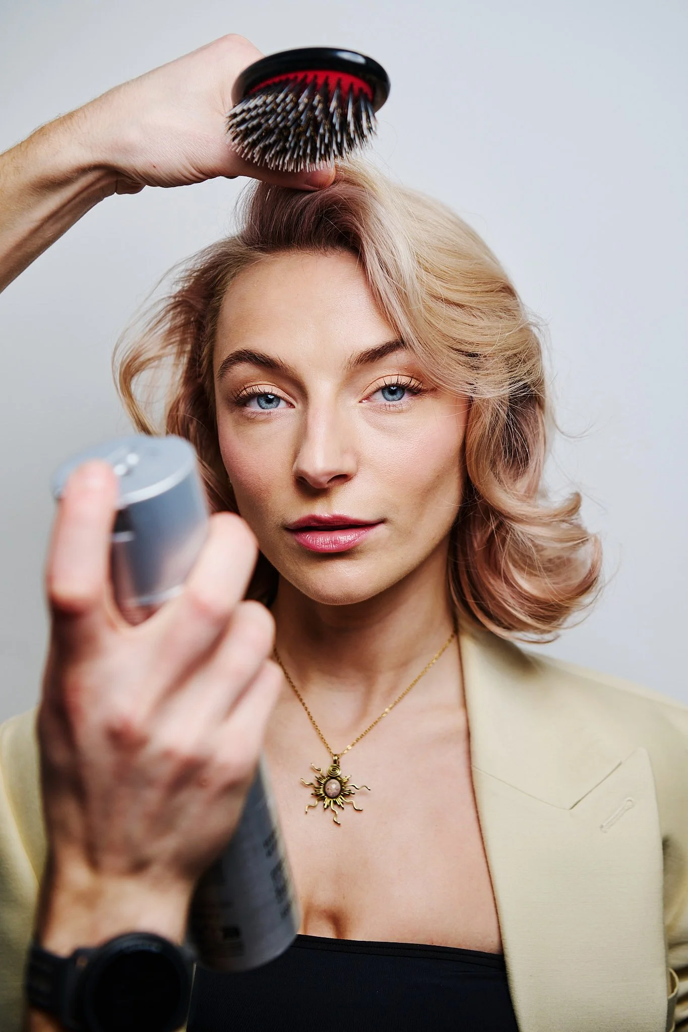 A woman with blonde hair getting her hair styled with a round brush by a stylist holding a can of hairspray.