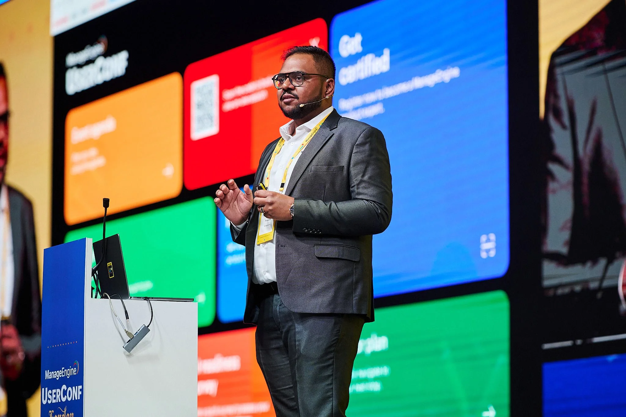 A man in a gray suit and glasses speaking at a conference with a large colorful screen behind him displaying various icons and text.