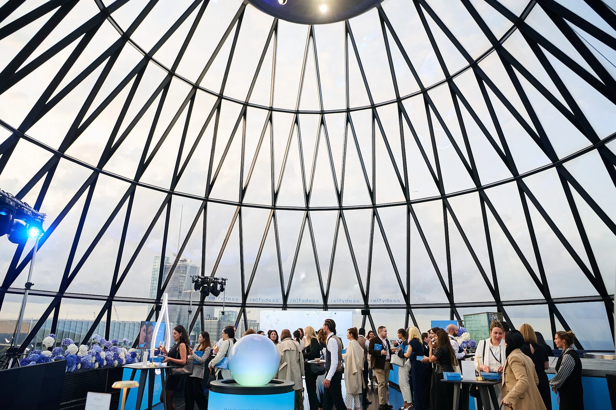 People gathered inside a modern, glass-domed structure at a conference or event, with a display of flowers and large screens in the background.