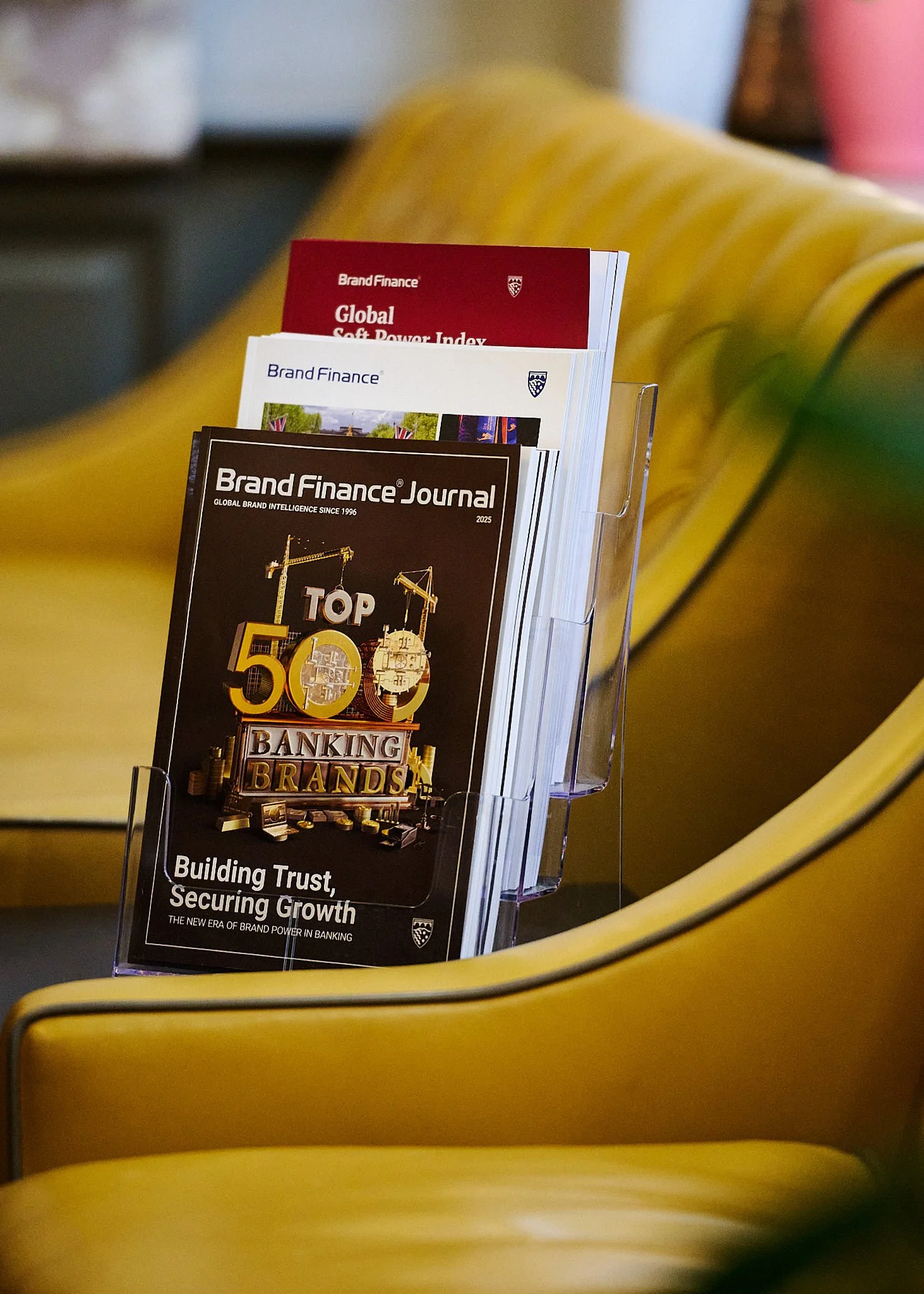 A clear plastic holder on a yellow chair contains several magazines, including one titled 'Top 50 Banking Brands' from Brand Finance Journal and others related to finance and branding, set against a blurred background.