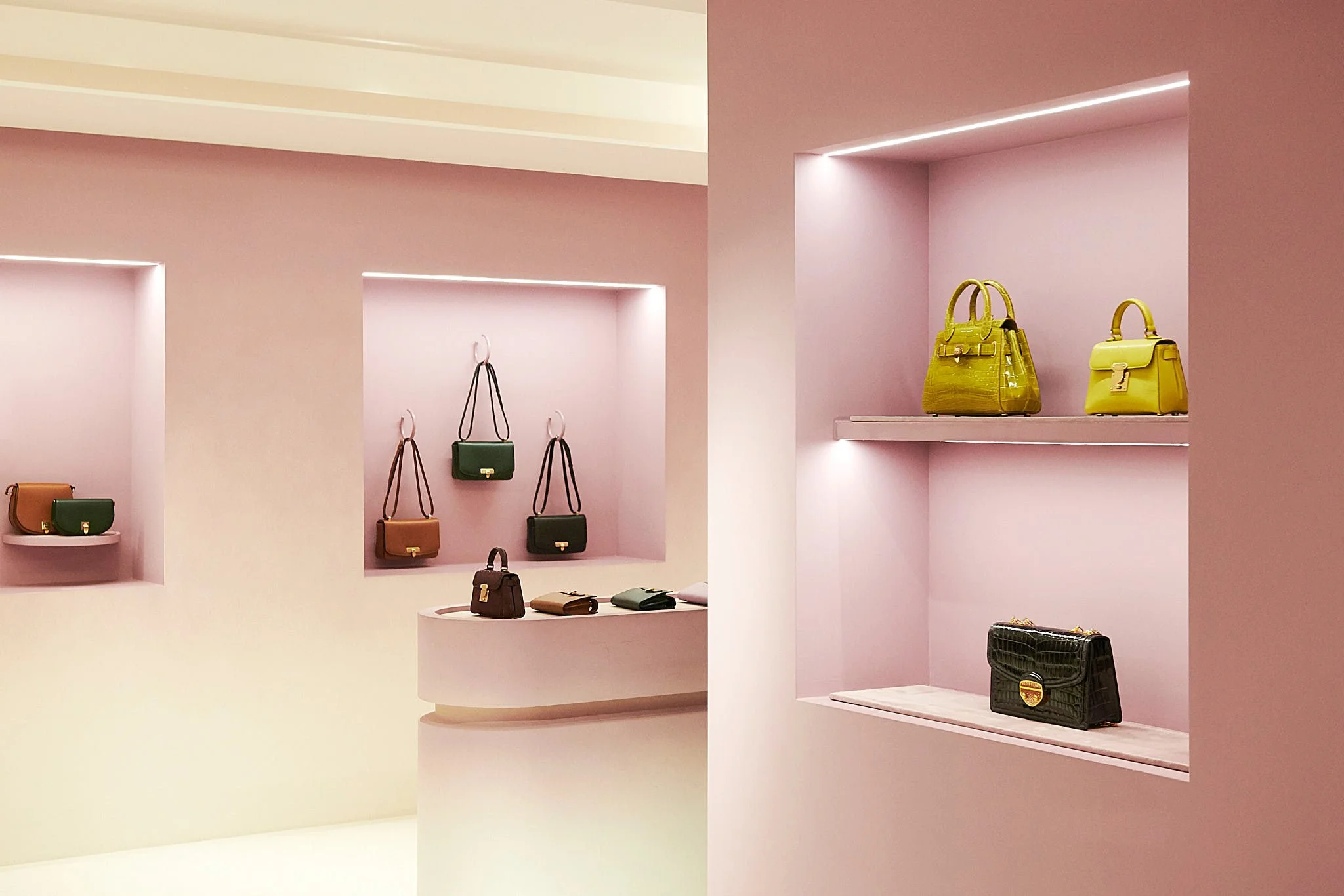 Display of designer handbags in a boutique with pink walls and shelves, illuminated by soft lighting.
