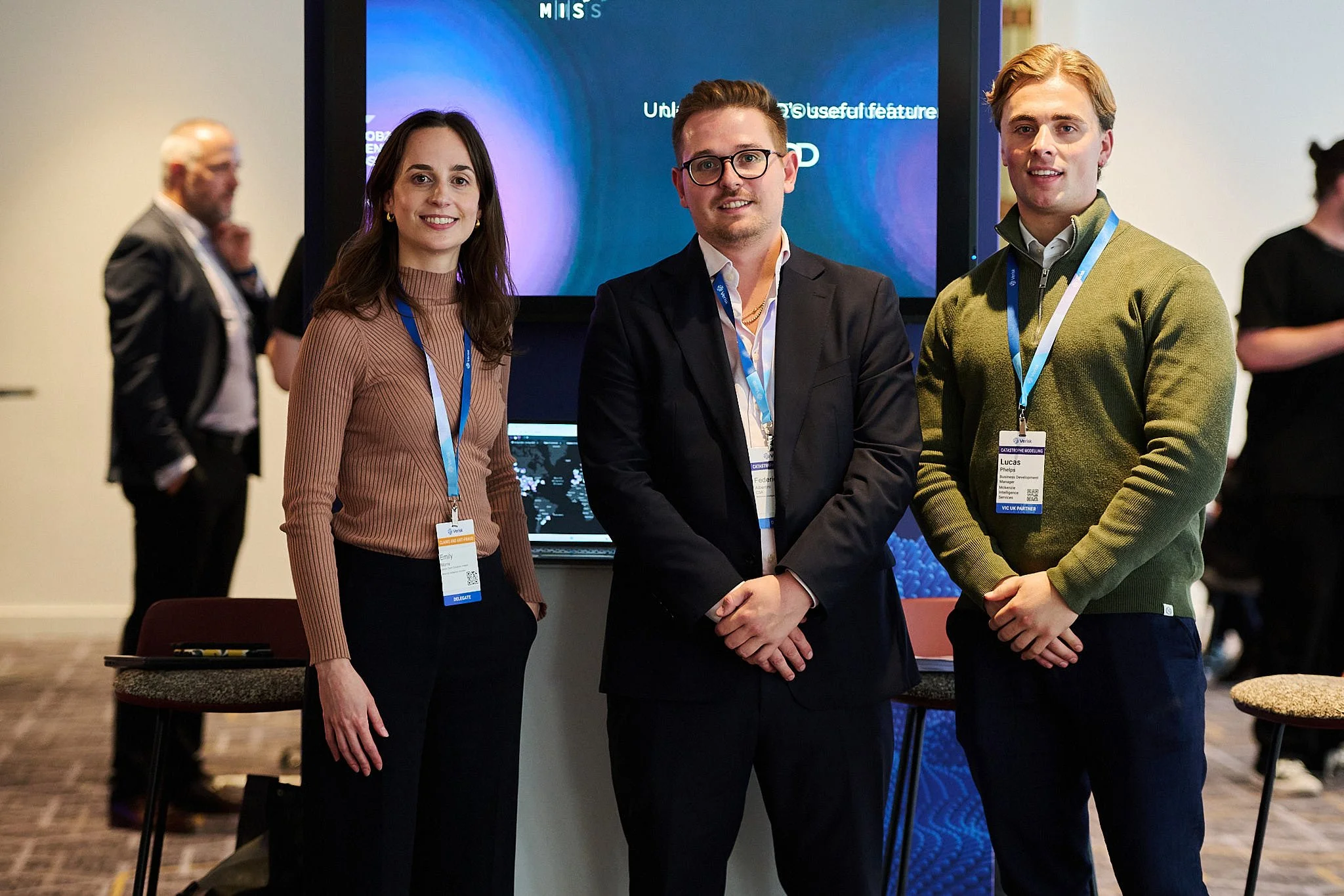Three people standing in front of a presentation screen at a conference, smiling at the camera. The woman on the left has brown hair, a brown ribbed sweater, and a name tag. The man in the middle has glasses, light brown hair, a dark suit, and a name