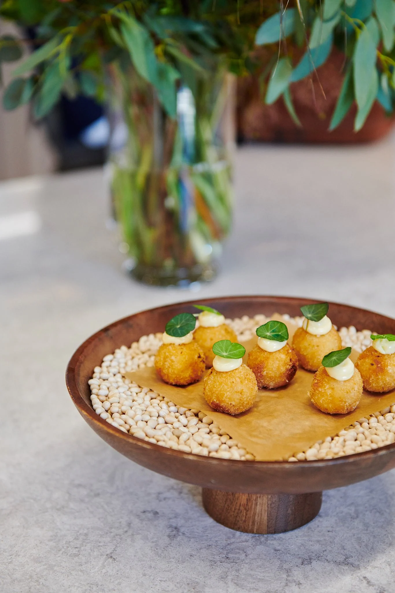 Plate of fried appetizers topped with cream and garnished with small green leaves, on a bed of white seeds, with a vase of green leafy plant in the background.