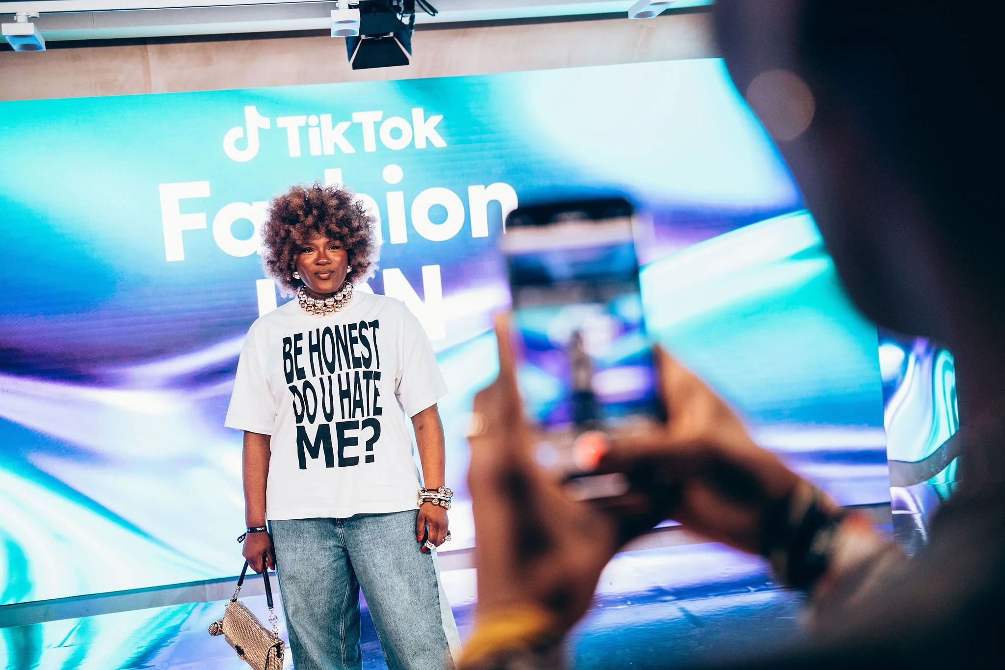 A woman with curly hair standing in front of a large screen displaying TikTok Fashion ICON. She is wearing a white t-shirt that says 'BE HONEST, DO U HATE ME?' and is holding a small handbag, with jewelry including a necklace and bracelets. Someone i