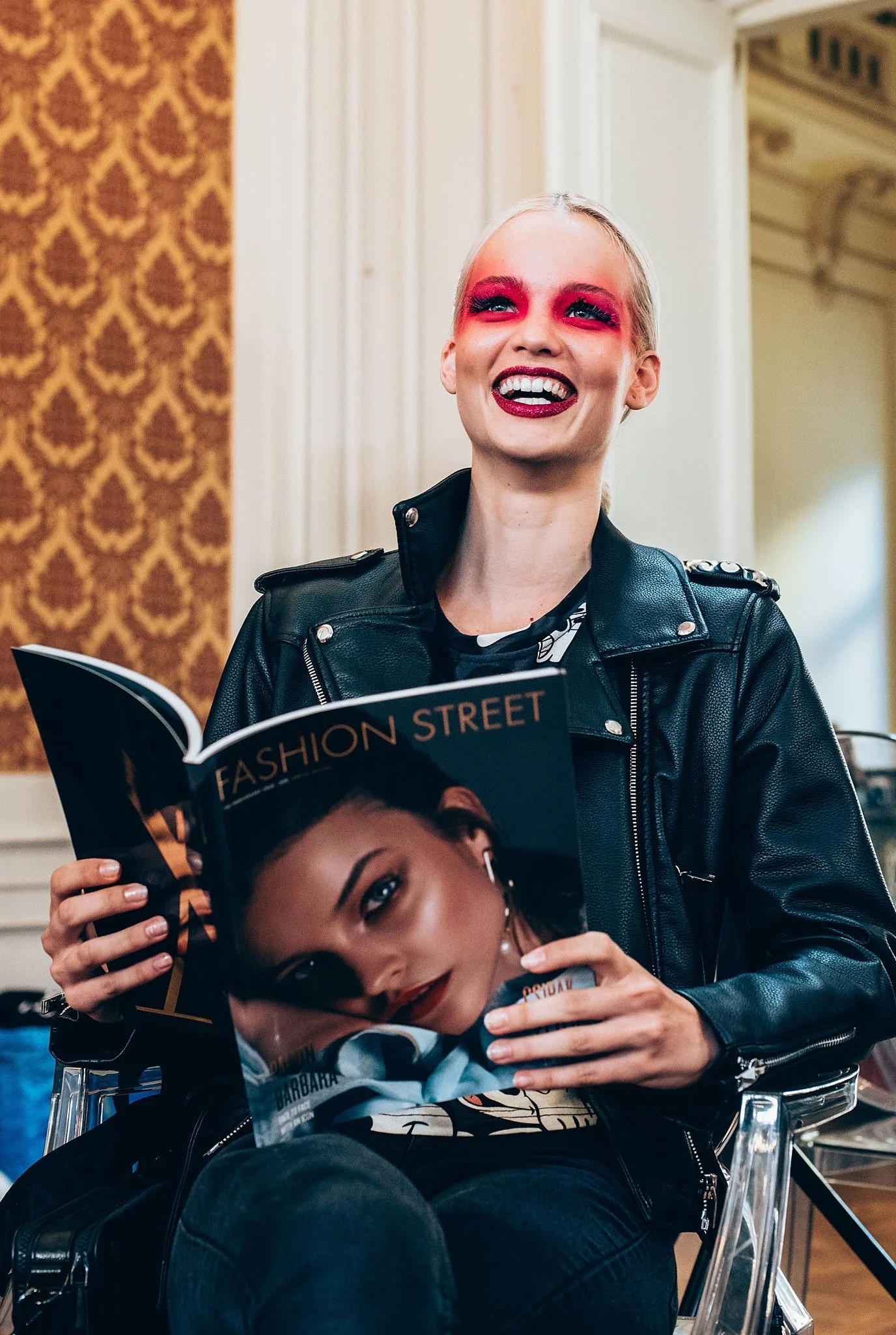 A young woman with blond hair and red makeup on her eyes and lips, smiling while holding a fashion magazine. She is wearing a black leather jacket and sitting on a transparent chair in an ornate room.