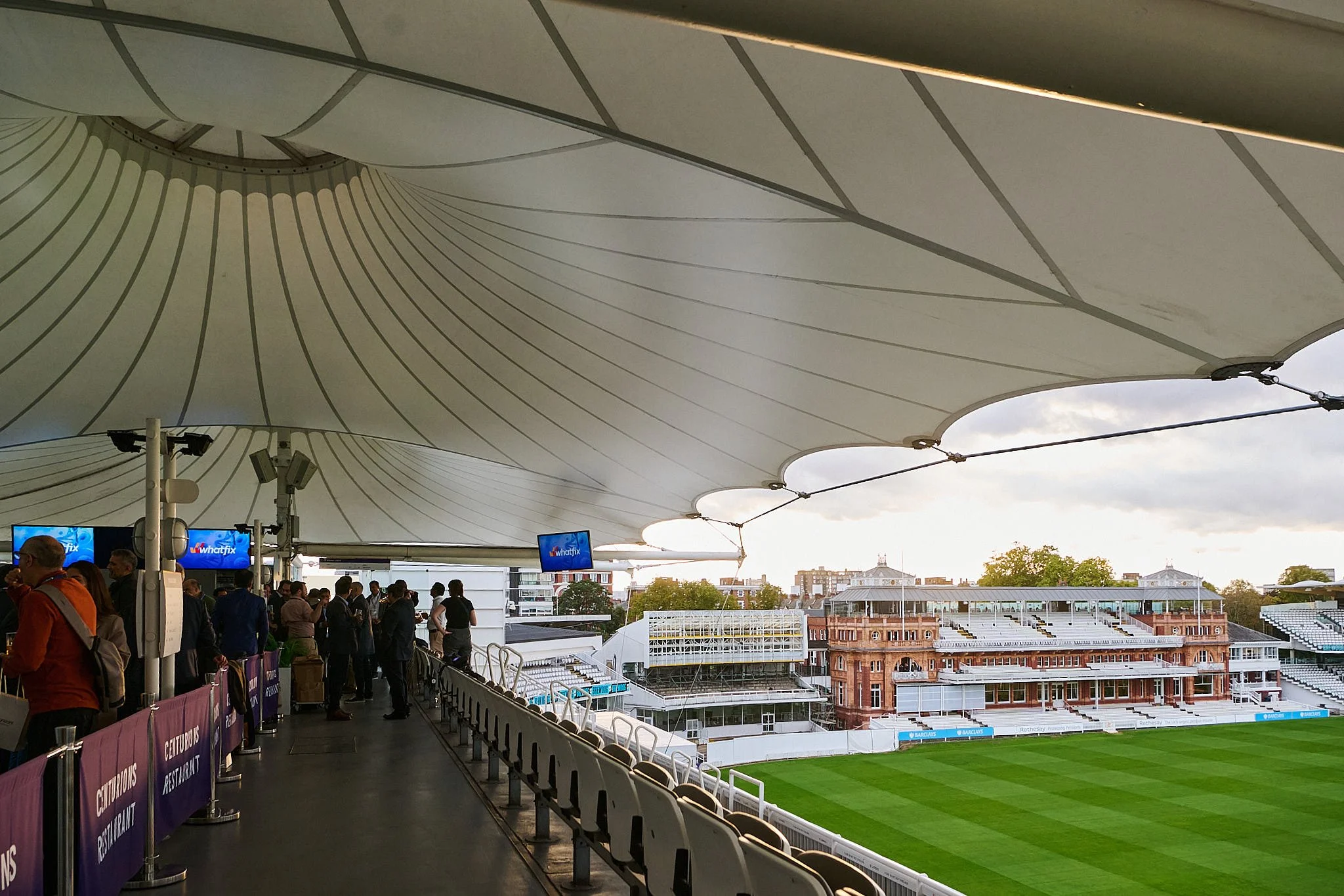 People waiting in line at a stadium concourse with a view of the field and surrounding buildings, under a large white canopy.