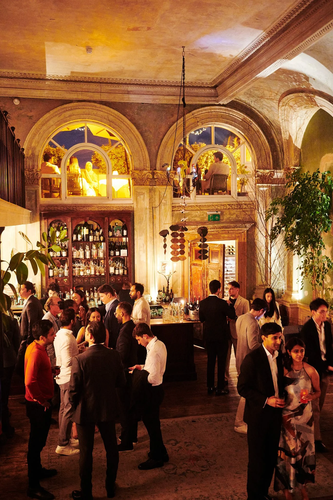 A lively indoor social gathering in a decorated establishment with people talking, holding drinks, and a bar area in the background. The setting features warm lighting, large arched windows, and ornate decor.