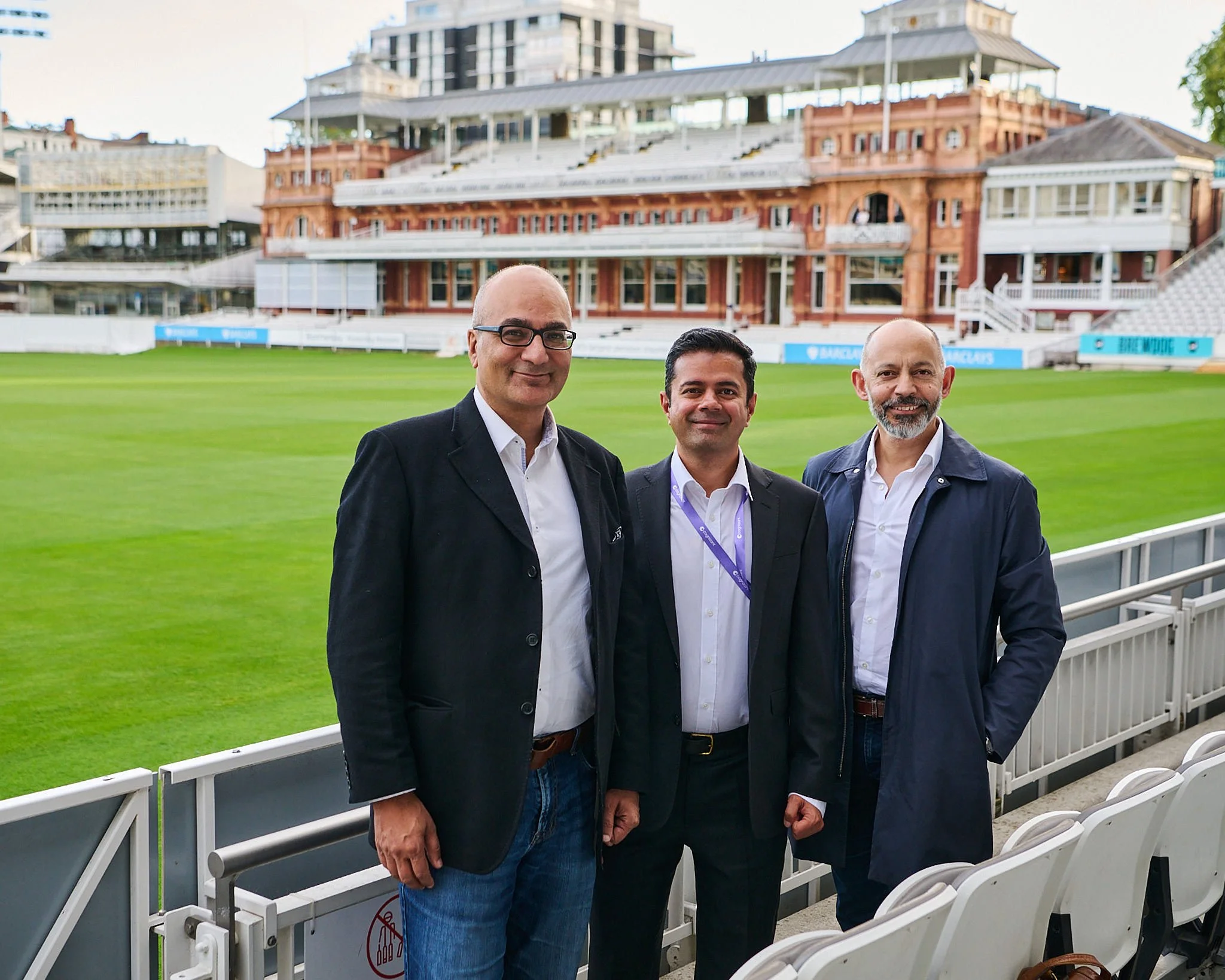 Three men in suits standing on a cricket stadium's sidelines with a lush green field and grandstand in the background.
