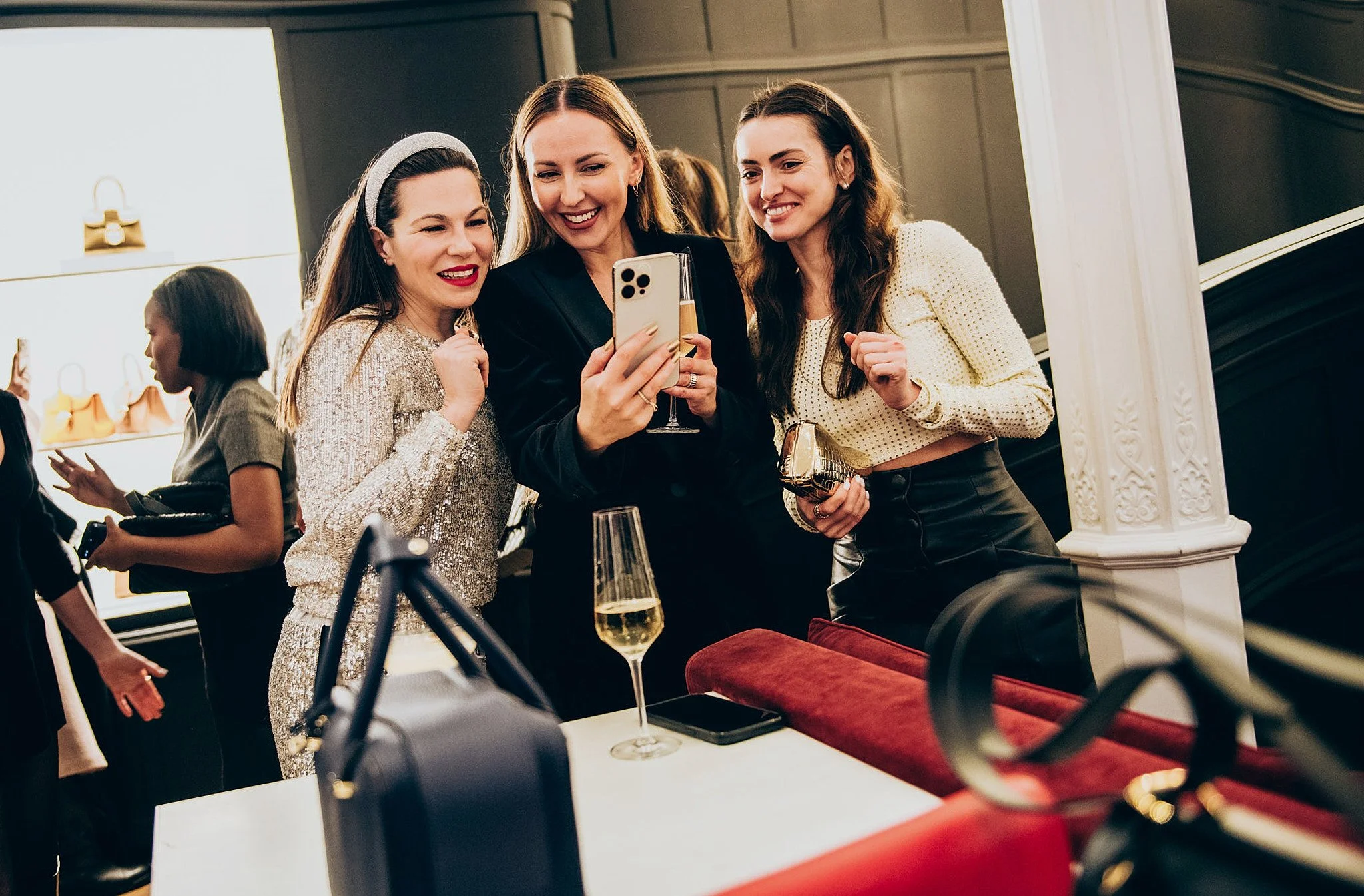 Three women in stylish outfits taking a selfie with a smartphone at a social gathering, with drinks and accessories on the table in a luxurious setting.