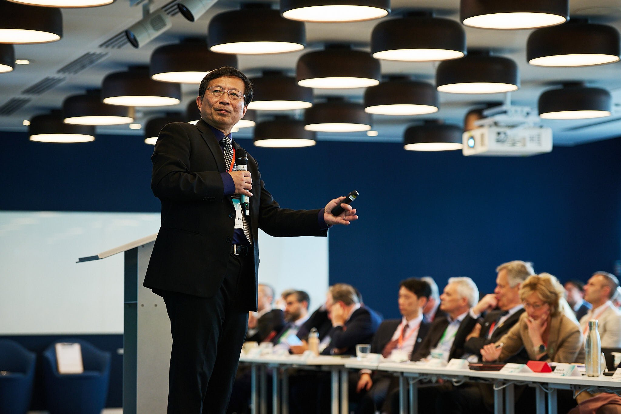 Man in a suit giving a presentation with a microphone in a conference room filled with seated audience members.