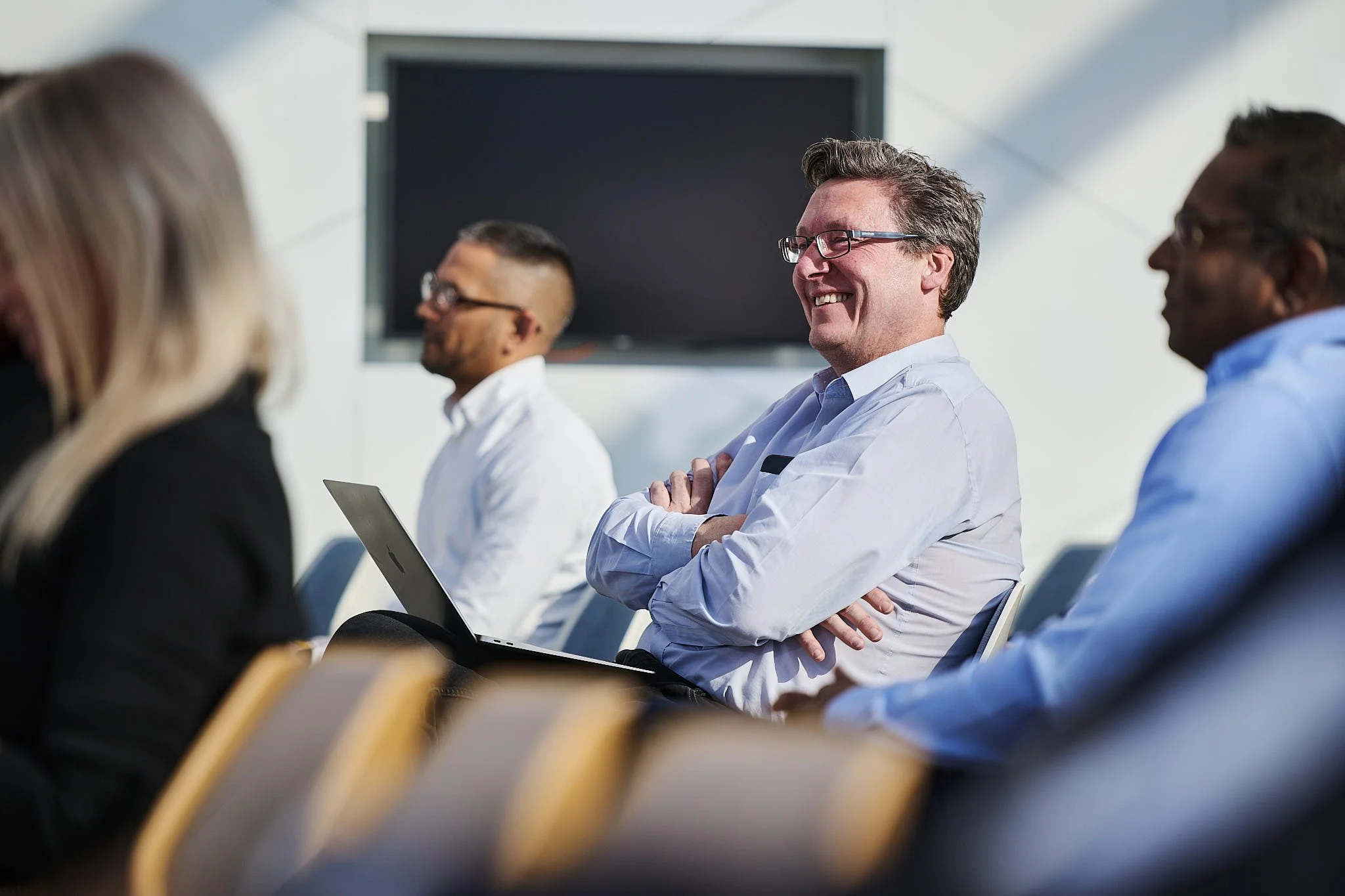 Business conference with professionals sitting in a row, some smiling and listening attentively, with a large screen behind them.