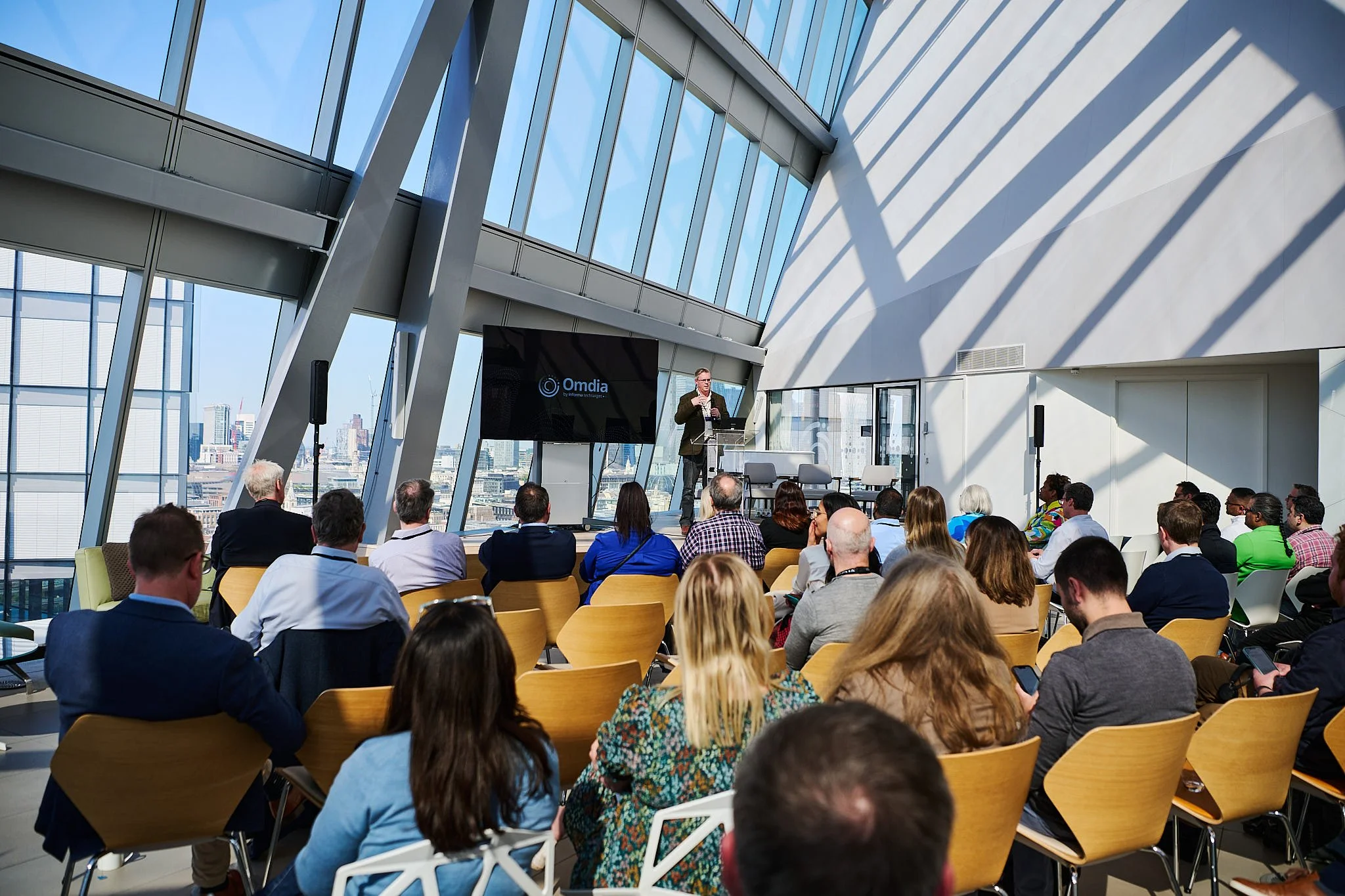 A diverse group of people attending a conference in a modern glass-walled room with a speaker on stage.