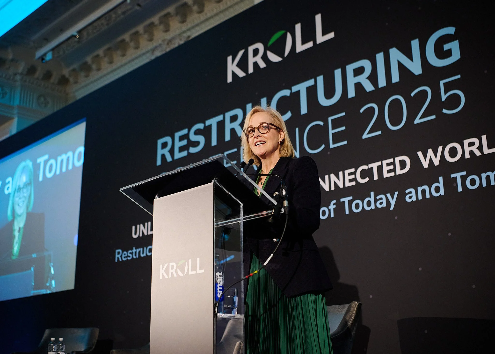 A woman with blonde hair and glasses speaking at a podium during a conference. Behind her is a large black screen with the words 'RESTRUCTURING' and '2025'.