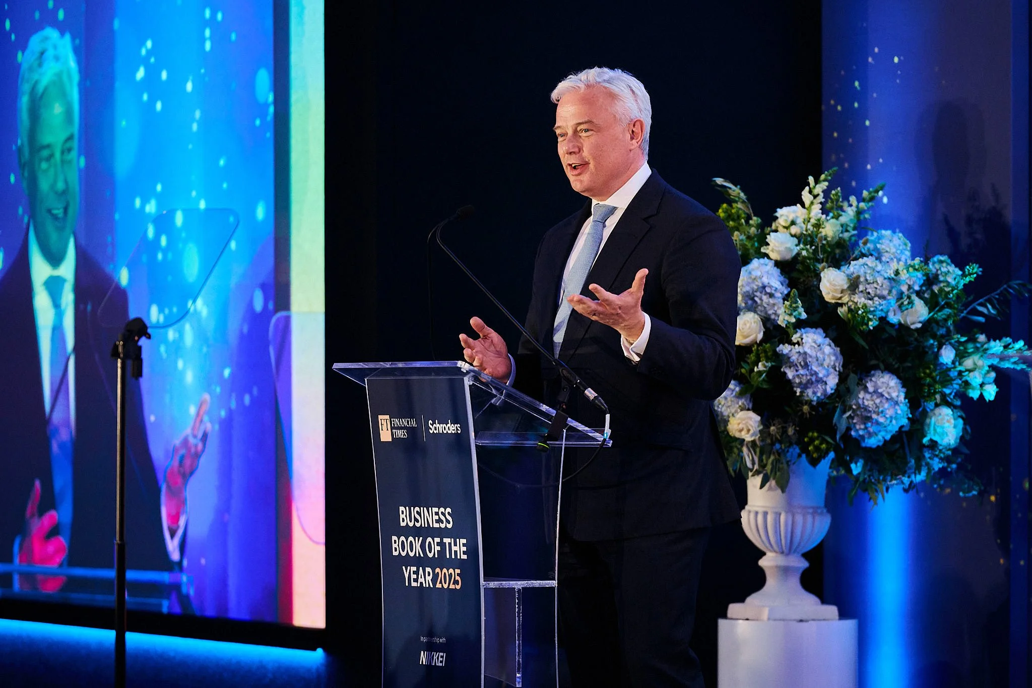 A man in a dark suit and light blue tie giving a speech at a podium during a formal event, with a large floral arrangement and a screen displaying his image in the background.