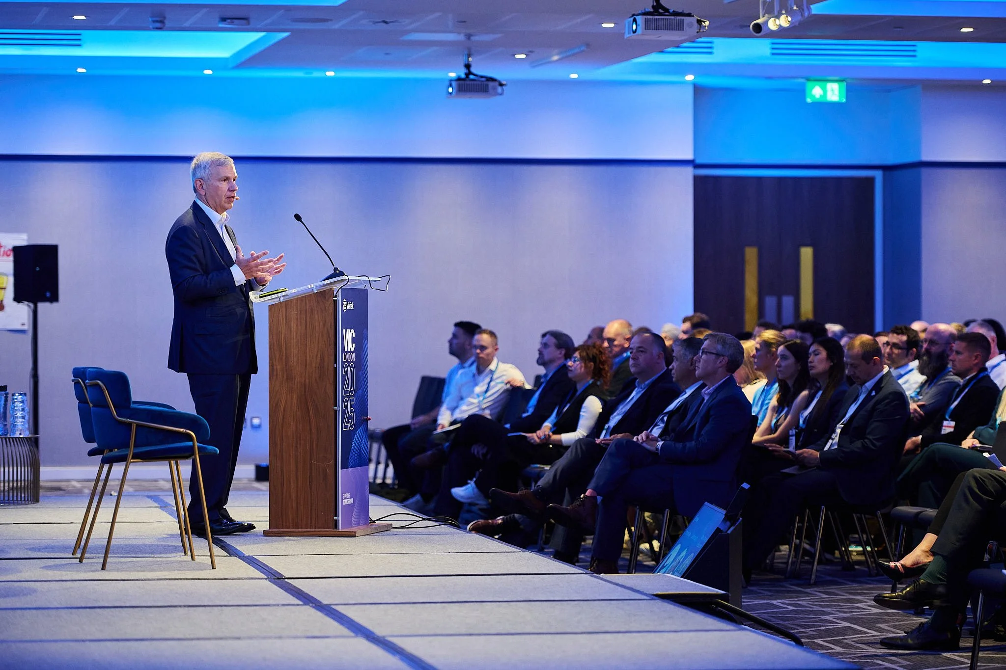 A man in a suit giving a presentation at a conference. The audience is seated and listening attentively in a dimly lit room with blue lighting.