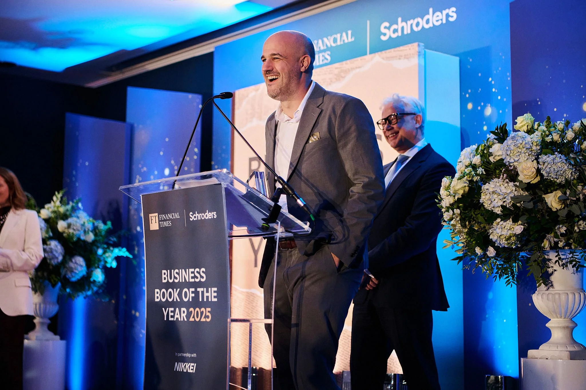 A man in a gray suit giving a speech at a podium during an awards event, with a smiling woman in glasses and a dark suit standing behind him. There are large floral arrangements and a blue-themed backdrop that reads "Financial Times Schroders Busines