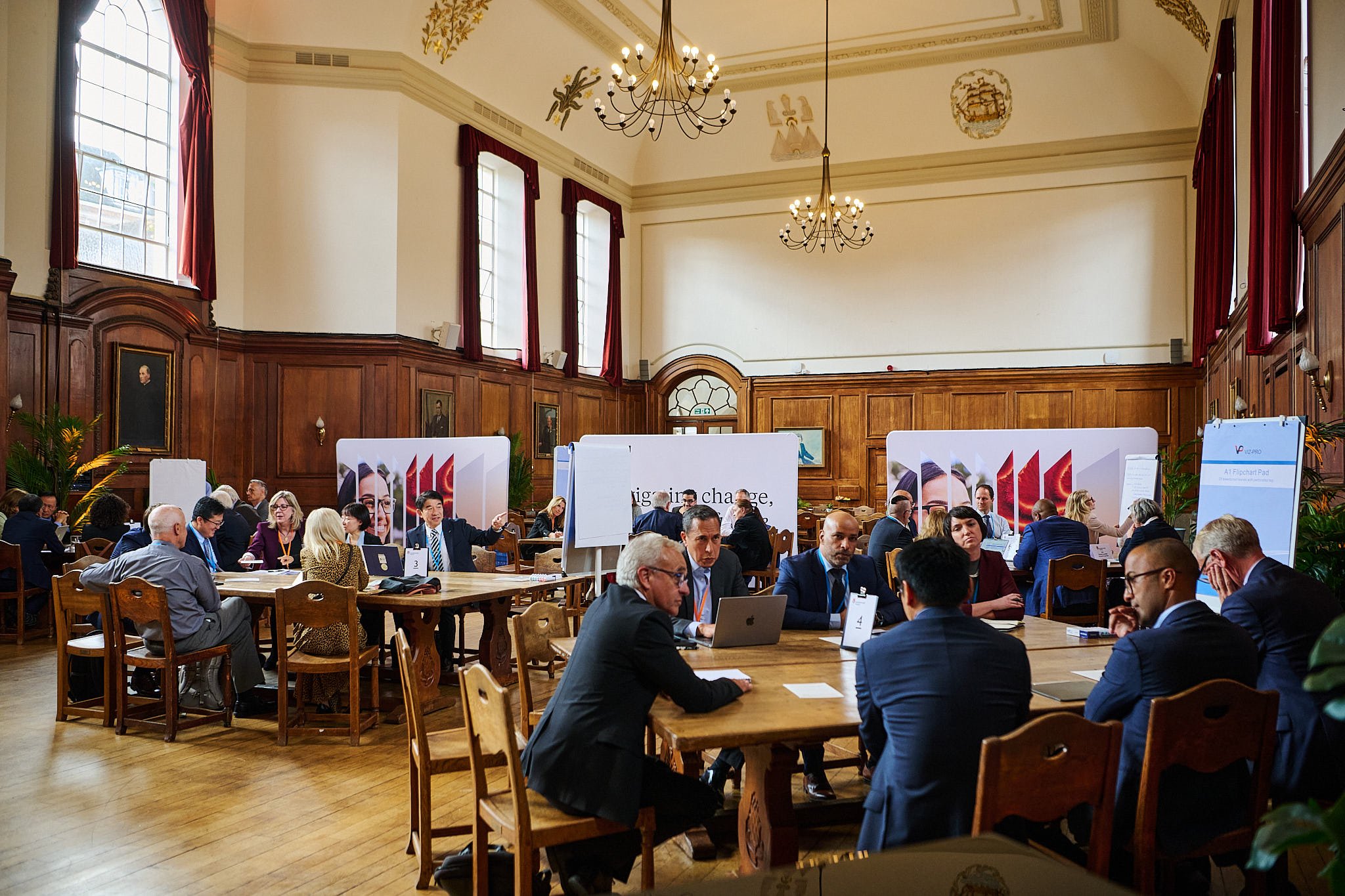 A large conference room with wood-paneled walls and tall windows with red curtains. Many people are seated at wooden tables, engaging in discussions. The room is decorated with chandeliers and portraits on the wall.