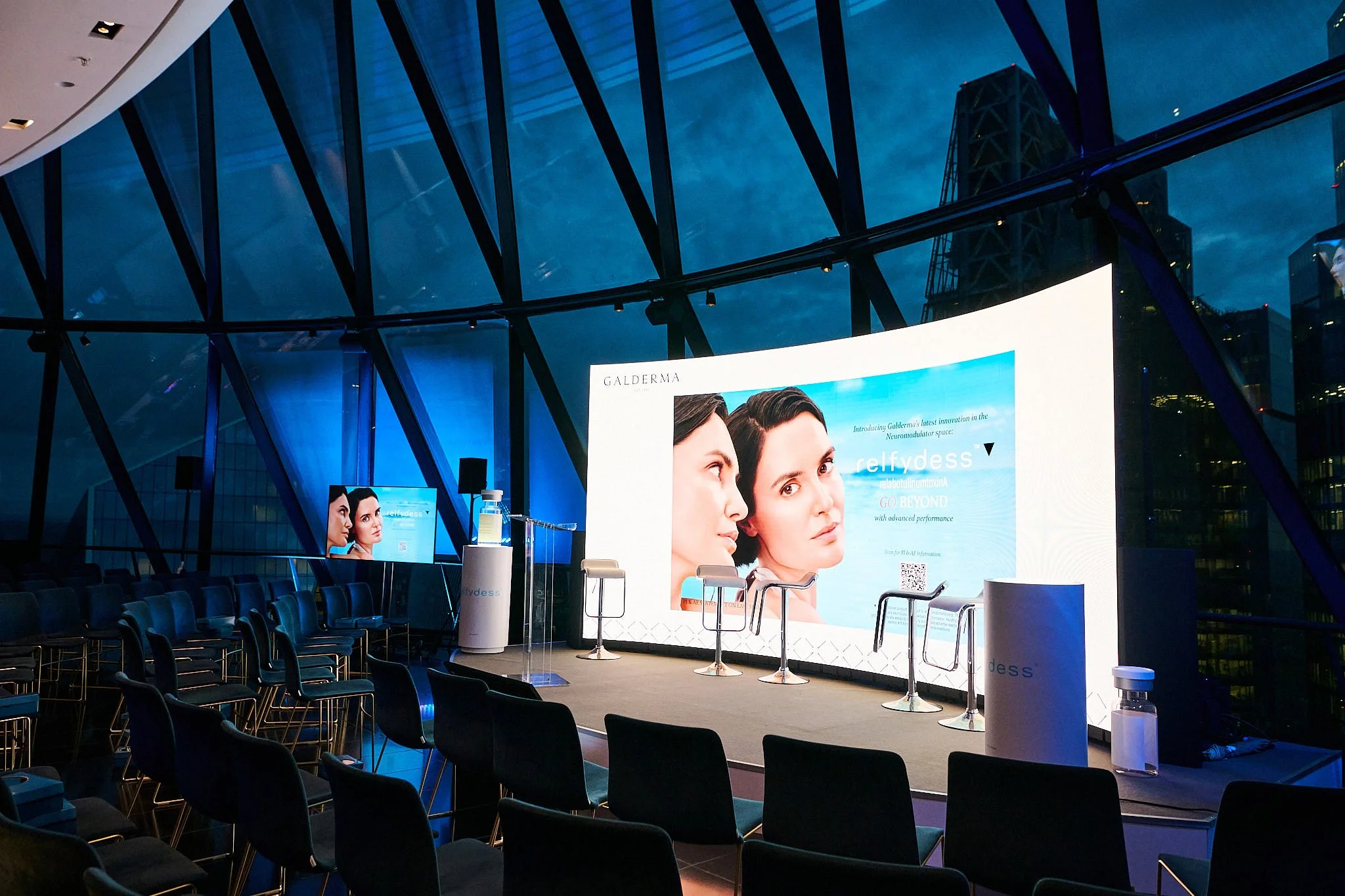 An indoor event space with a large digital screen displaying an advertisement for Galderma's skincare product, Selfydess, showing two women's faces in profile. There are several empty chairs arranged in rows facing the stage, and the space has floor-
