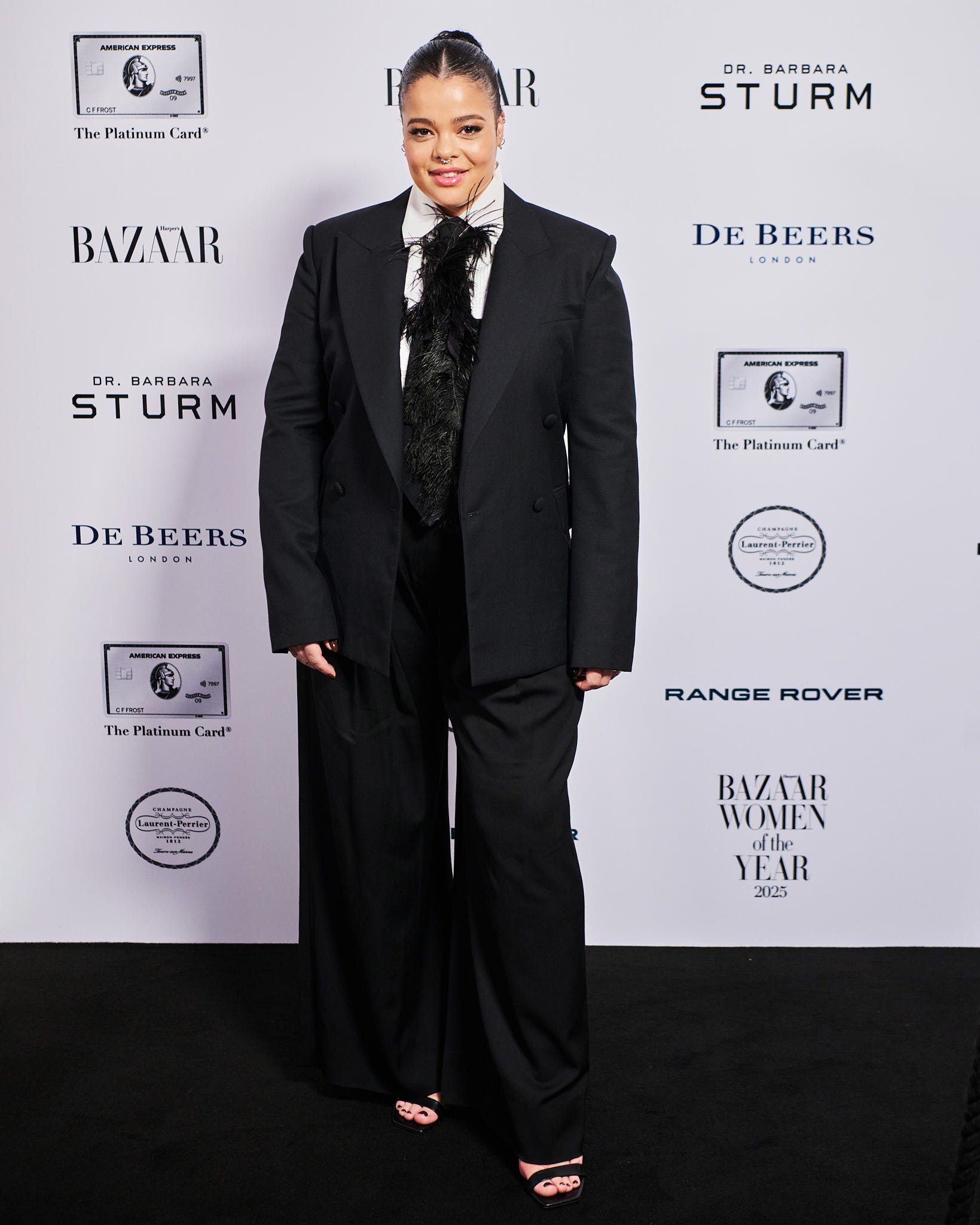 Woman in black suit standing on black carpet at event, backdrop featuring brand logos and event name.