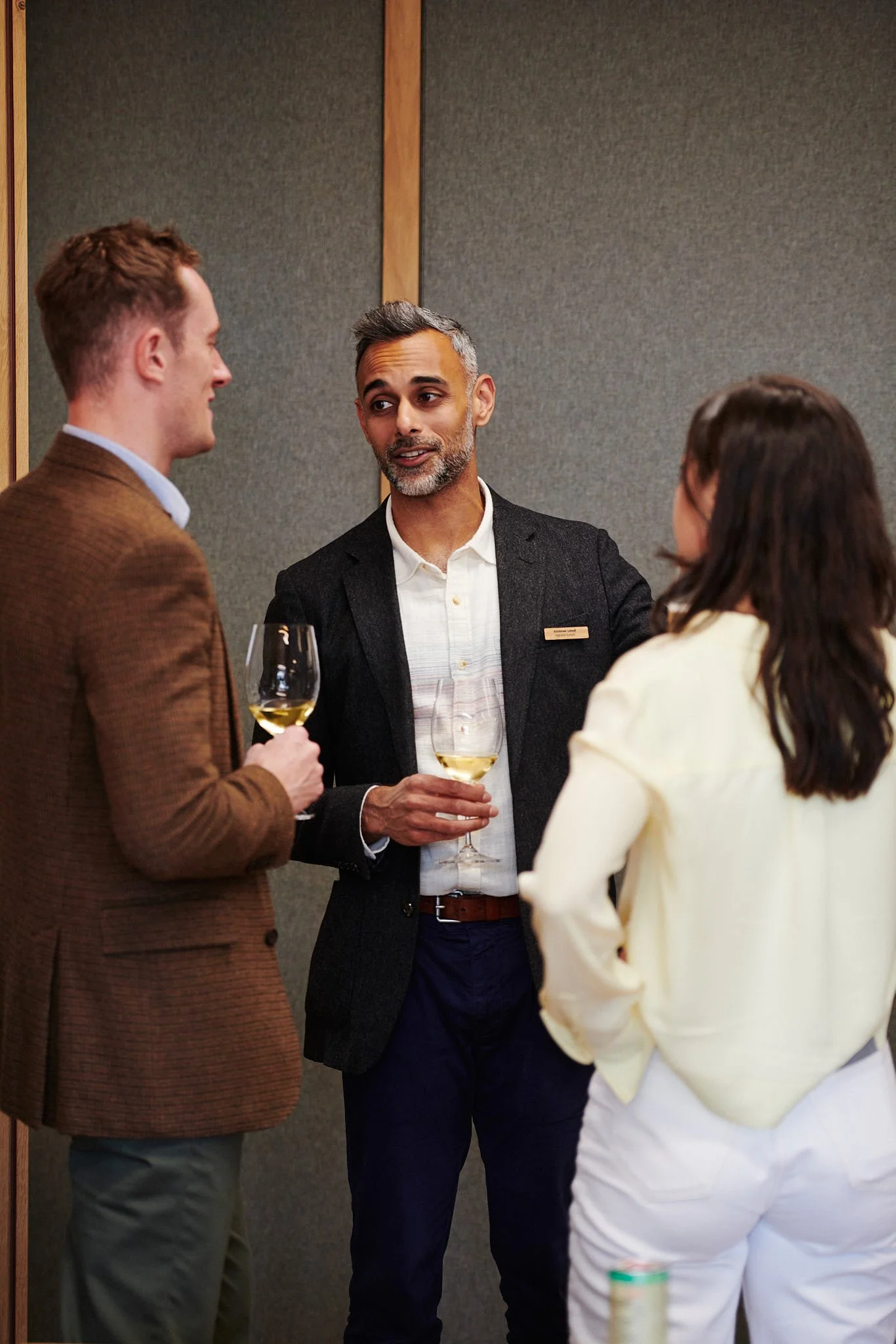 Three people having a conversation at a social event, each holding a glass of white wine.