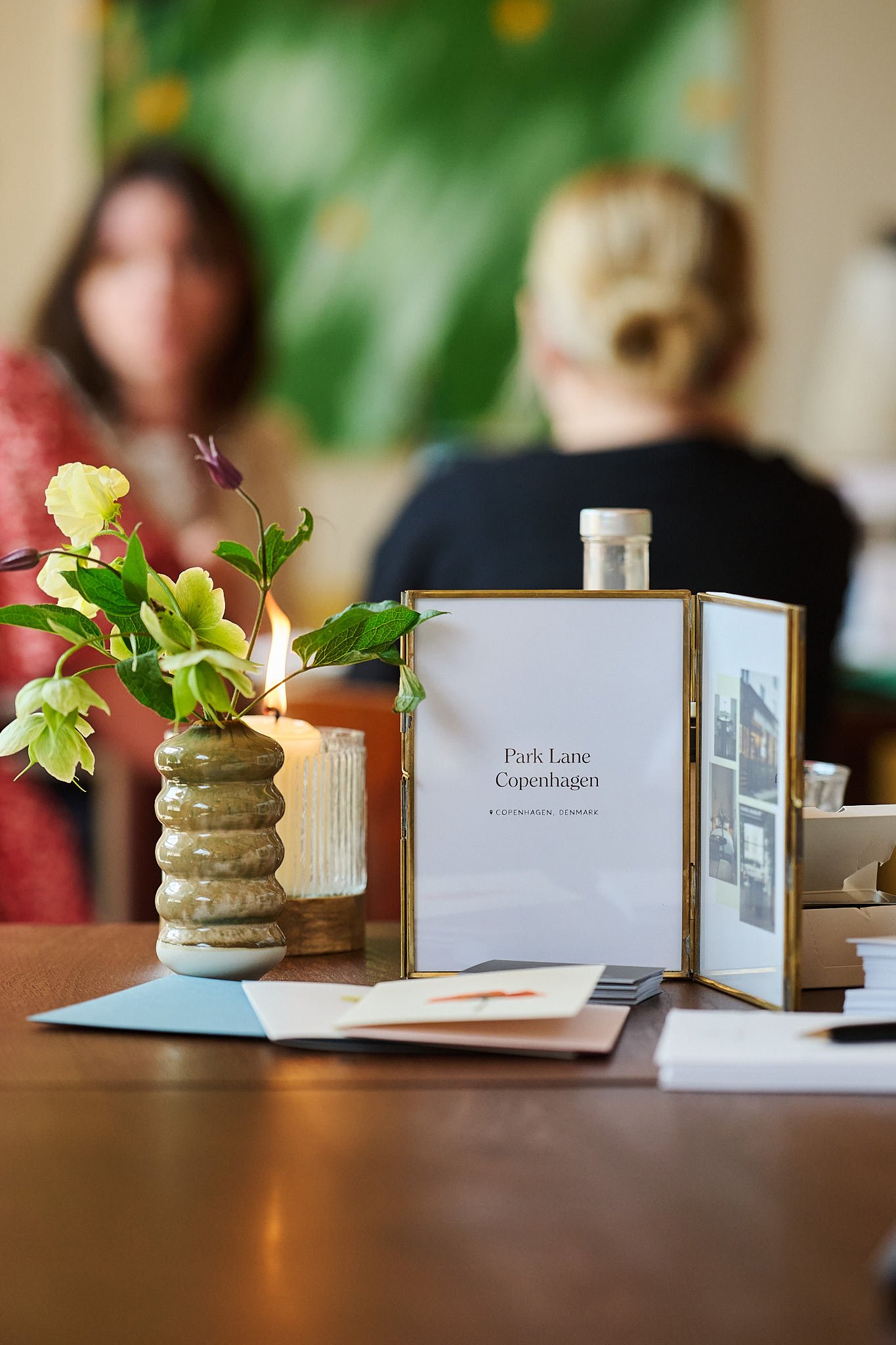 A dining table with a ceramic vase holding green flowers, a lit candle, and a framed sign reading 'Park Lane Copenhagen, Denmark' with photographs inside the frame. In the background, two women are talking, one with blurred face and another with her 