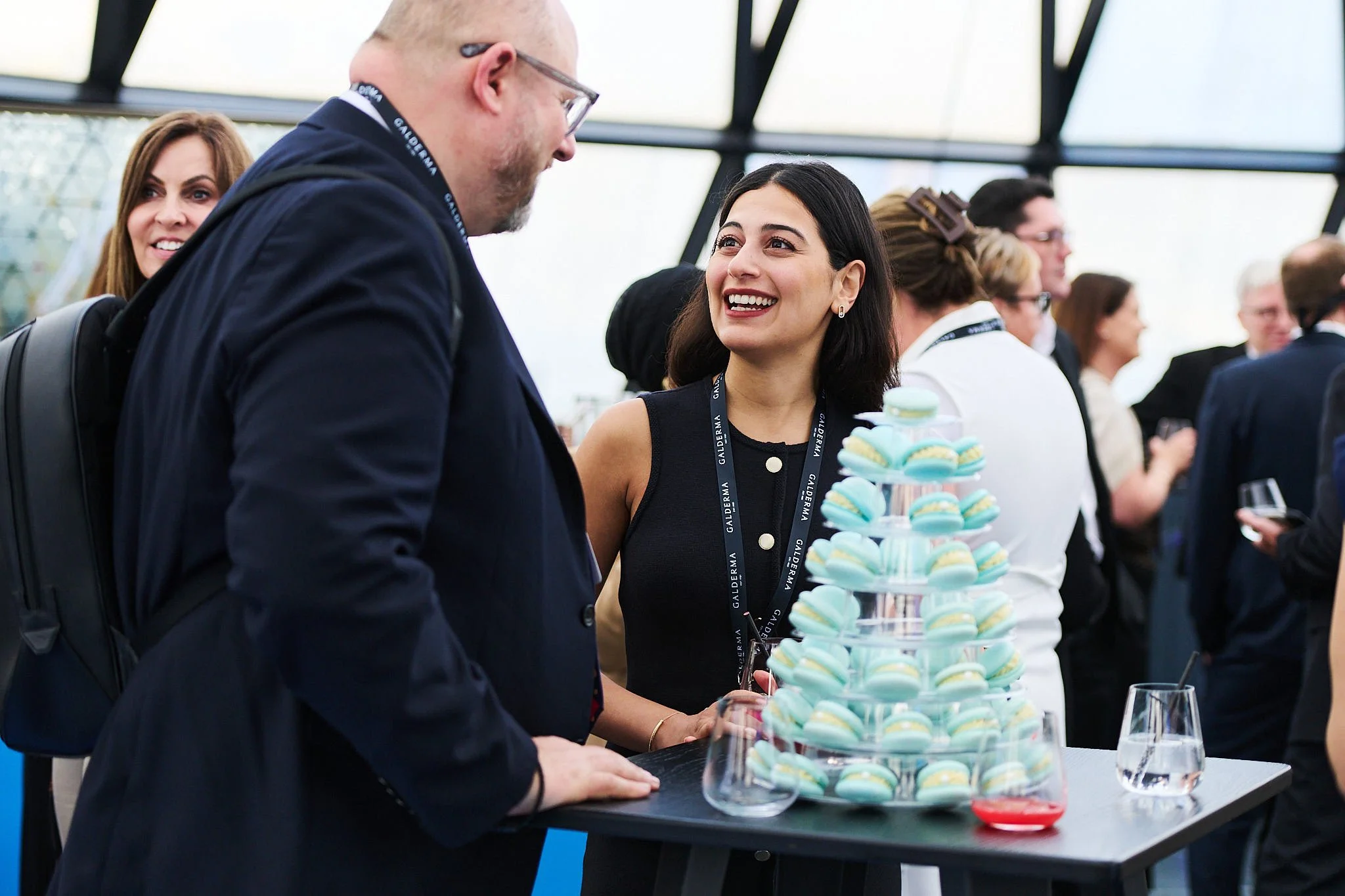 A woman in a sleeveless black dress and a man in a suit are talking at a table with a tiered platter of blue and yellow macarons during a professional event. Other people are mingling in the background.