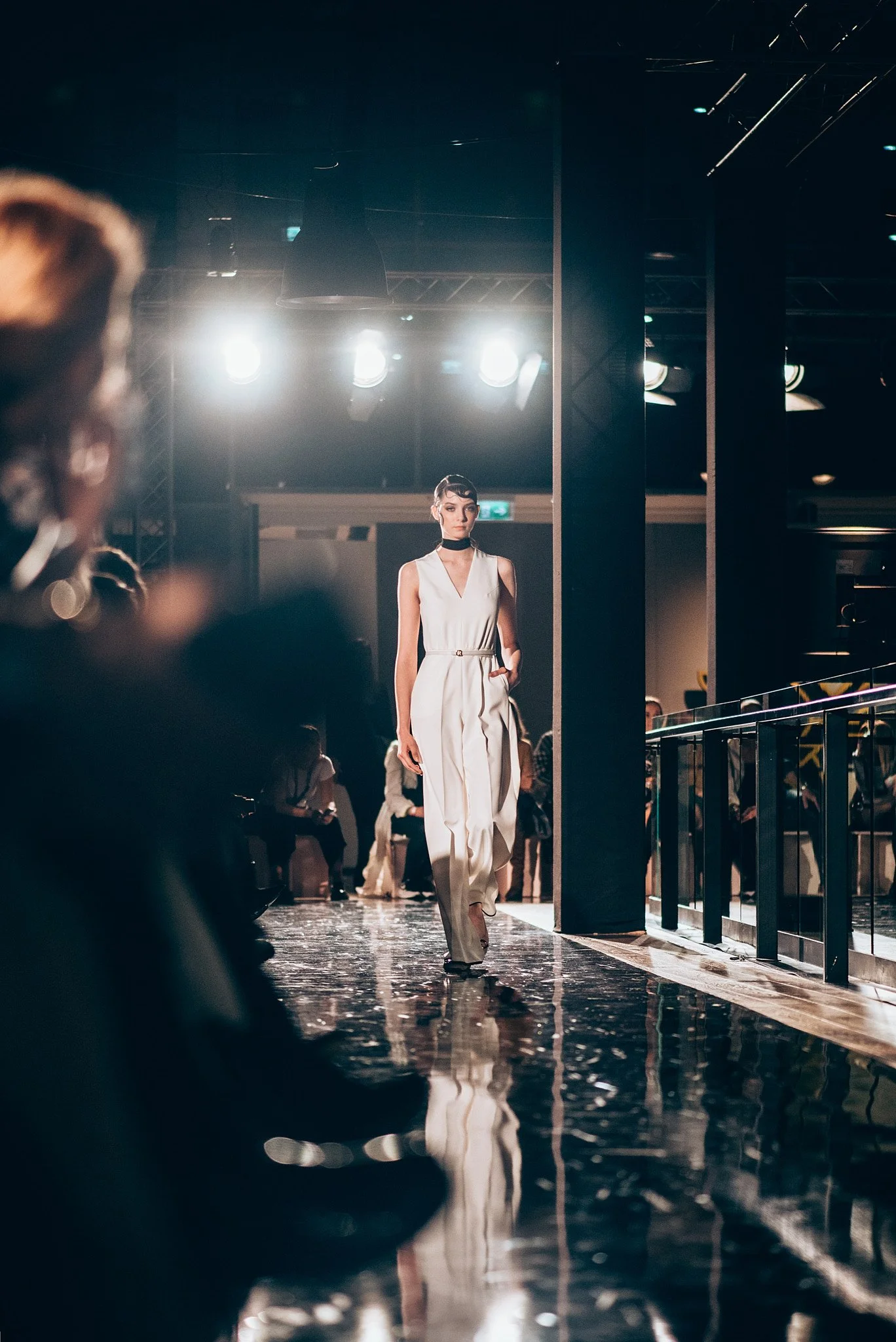 Model walking down the runway at a fashion show, illuminated by bright lights, wearing a sleeveless white jumpsuit with a belt and black choker, with an audience watching.