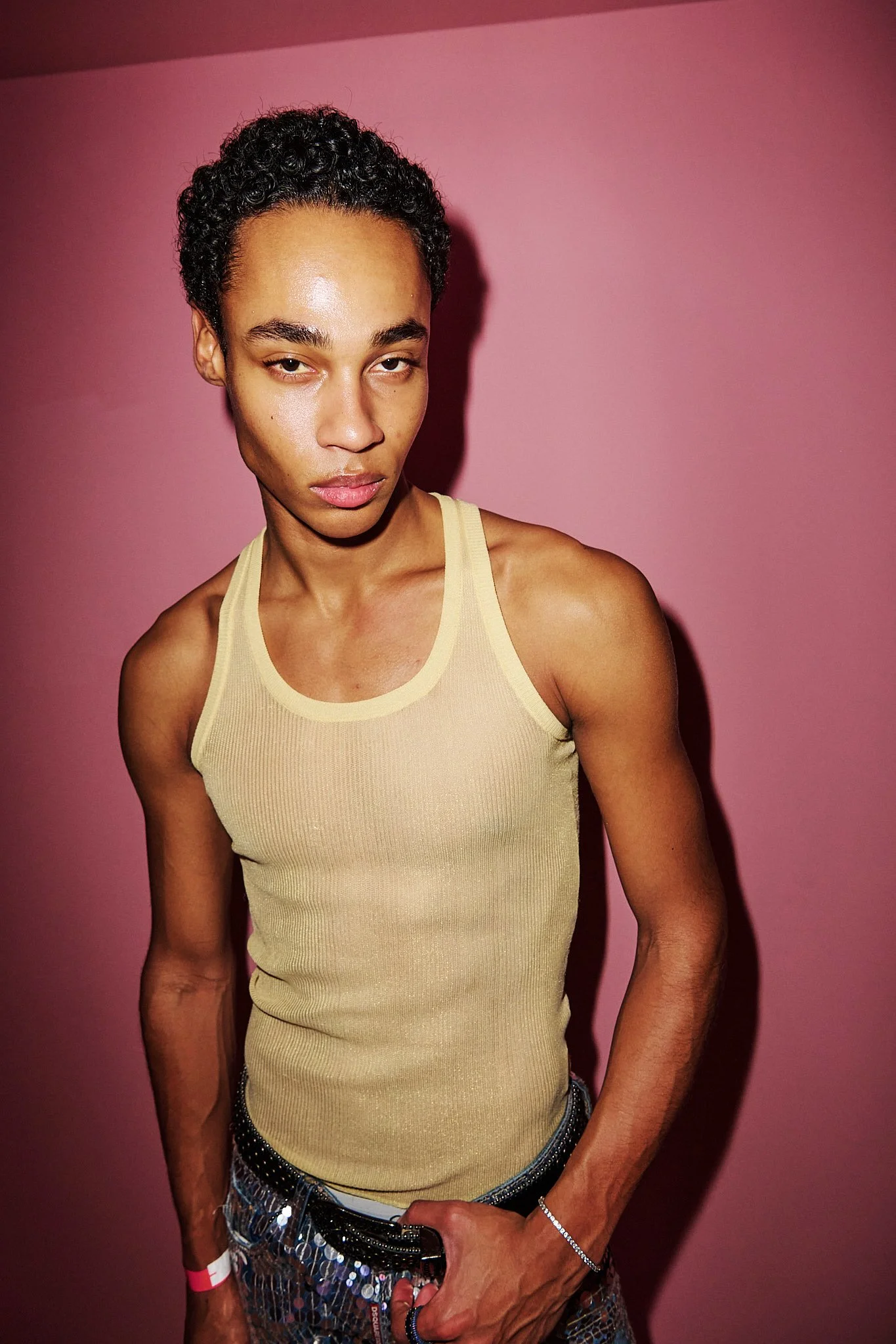 A person with short, curly black hair wearing a beige tank top stands against a pink wall, looking at the camera with a slightly serious expression.