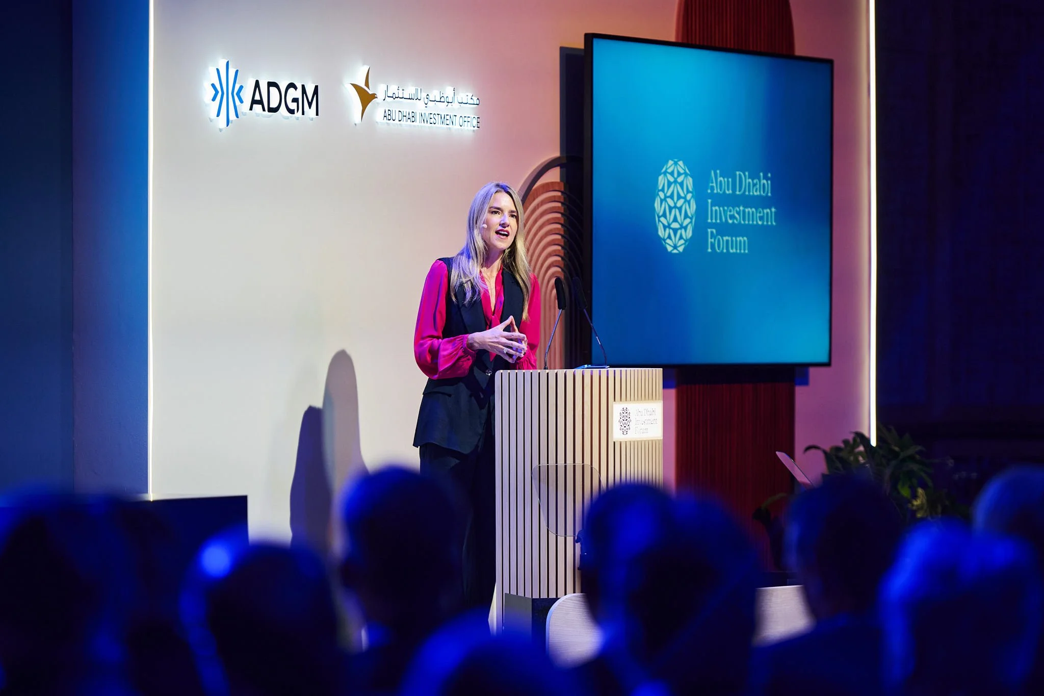 A woman in a pink blouse and black vest is speaking at a podium during a conference, with a large screen displaying the Abu Dhabi Investment Forum logo behind her and a white wall with logos and text to her left.