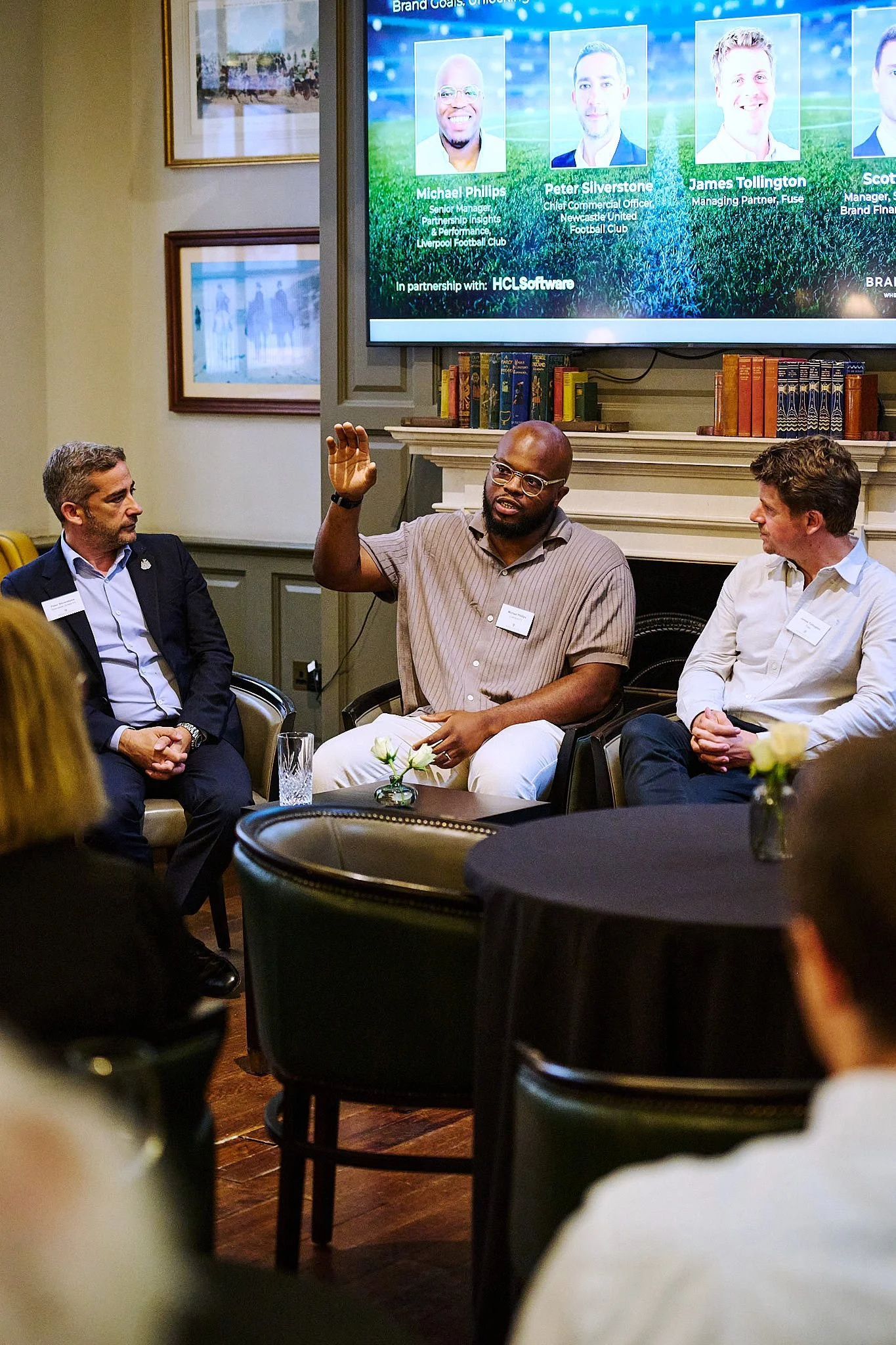 A man in a striped shirt and glasses is speaking and raising his hand during a panel discussion with two other men, one in a dark suit and the other in a white shirt. They are seated in a room with a large screen displaying speakers' names and titles