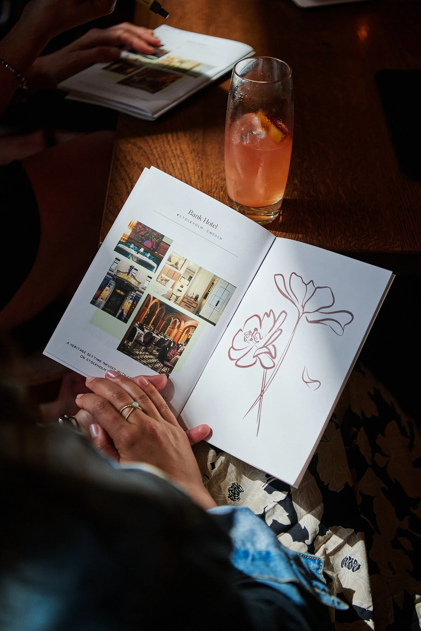 A person holding an open brochure showing images of the Bank Hotel in Stockholm, Sweden, with a floral line drawing on the right page. In the background, there is a glass with a pink-colored drink containing ice and a lemon slice, on a wooden table. Another person is seen browsing a magazine in the background.