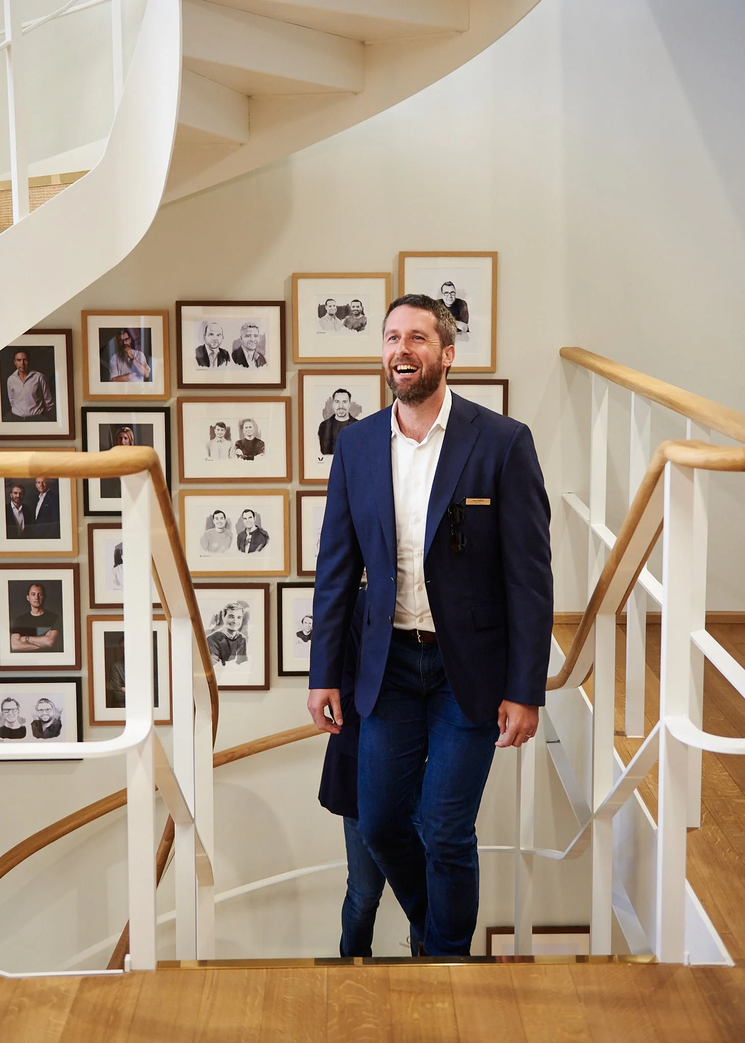 A man in a blue blazer and white shirt standing on a staircase with framed photos on the wall behind him, smiling and looking to his left.