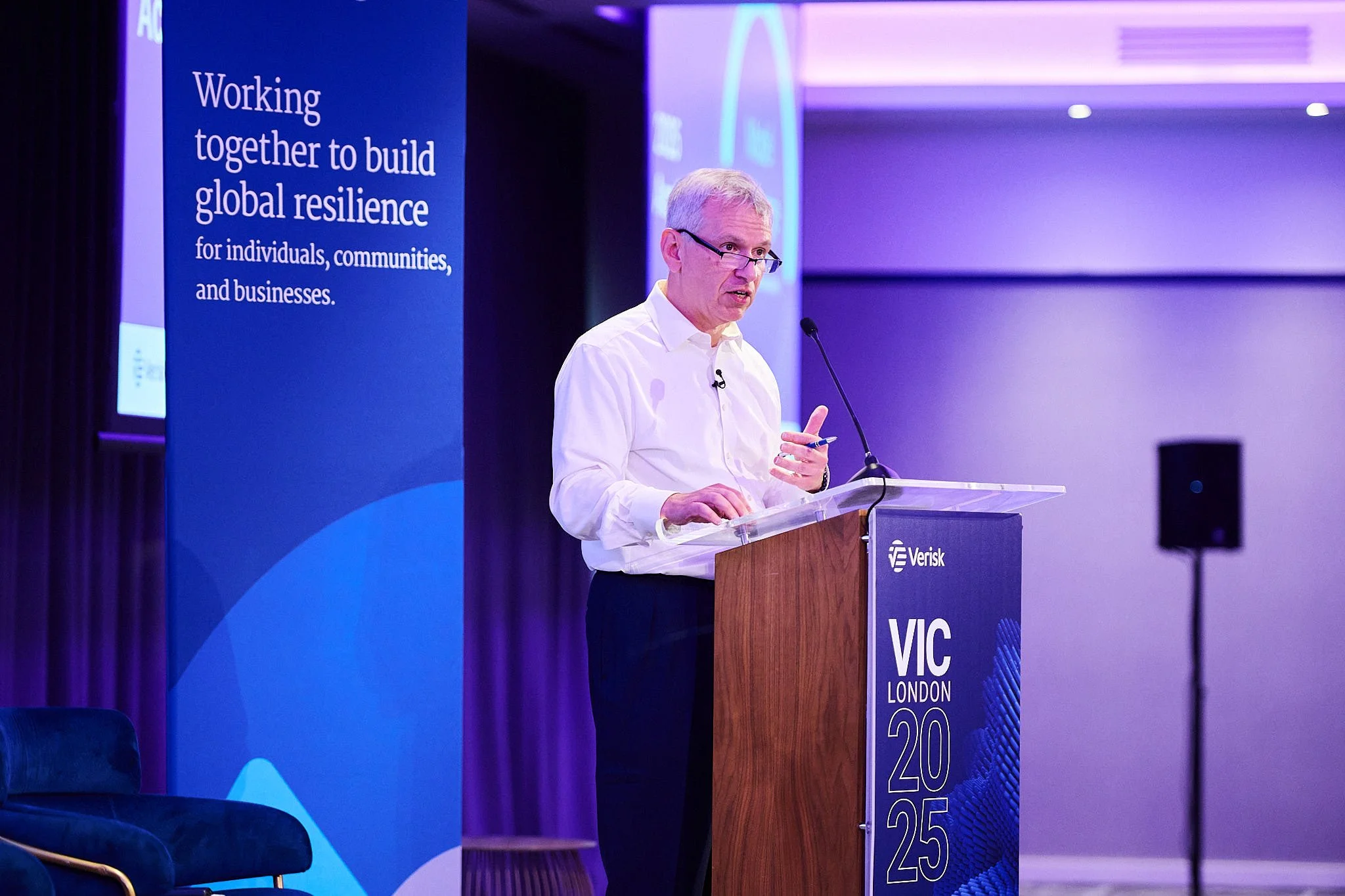 A man in a white shirt giving a presentation at a podium with a blue banner that reads "Working together to build global resilience for individuals, communities, and businesses." The event is titled "VIC London 2025."