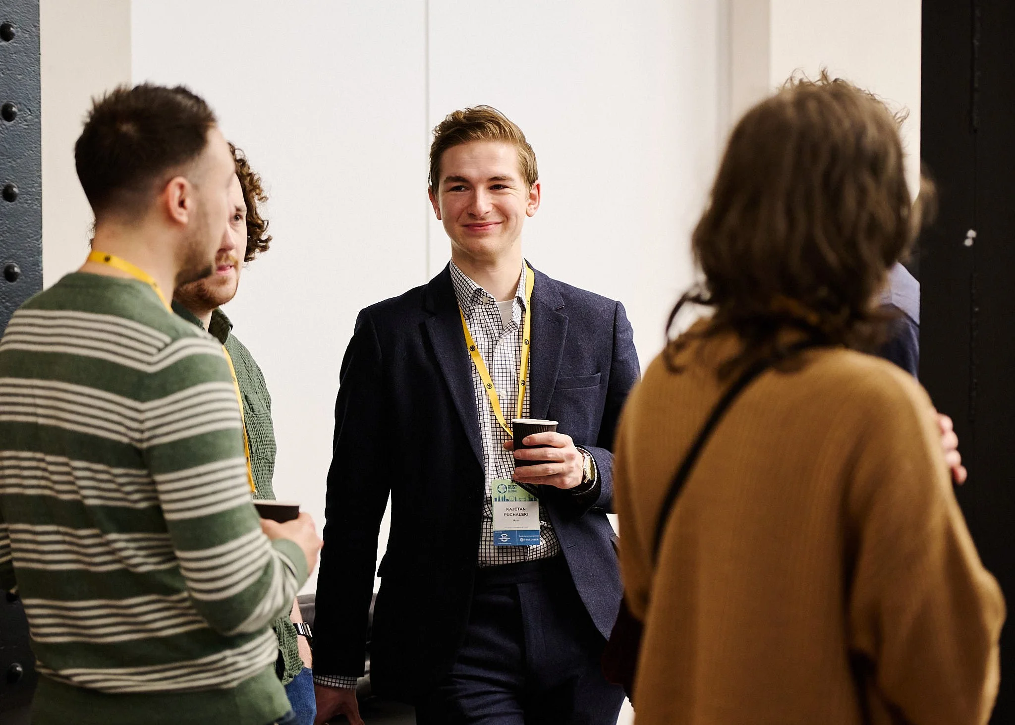 Group of young adults at a networking event, engaging in conversation, with one man smiling while holding a coffee cup.