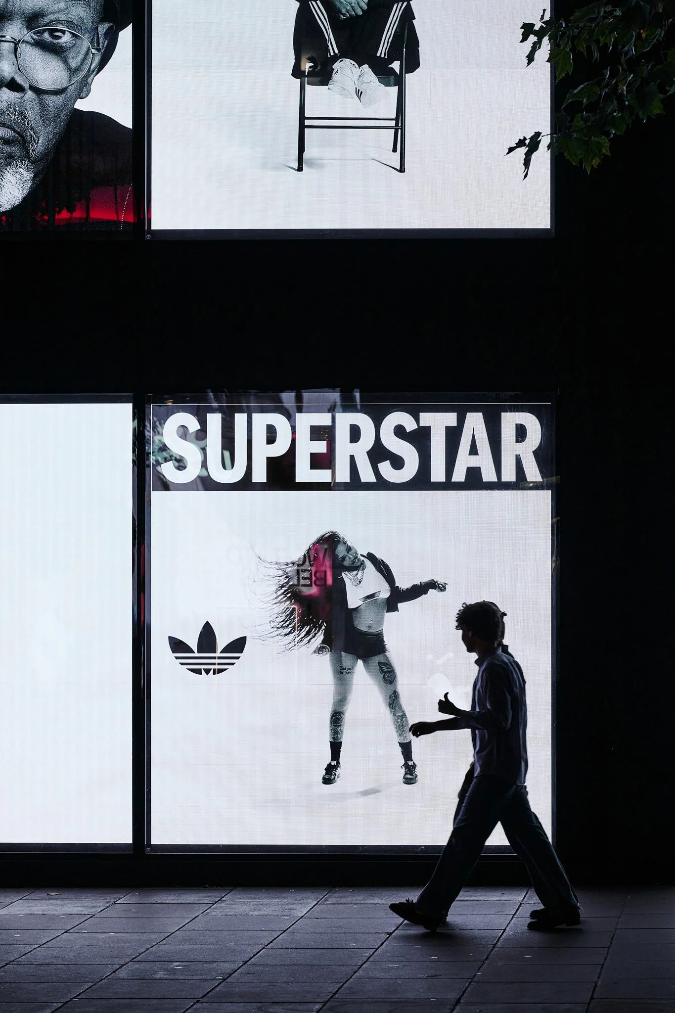 A large illuminated advertisement for Adidas featuring a woman dancing with a skateboard, the word 'SUPERSTAR' in bold white letters above her, and a person walking past in silhouette.