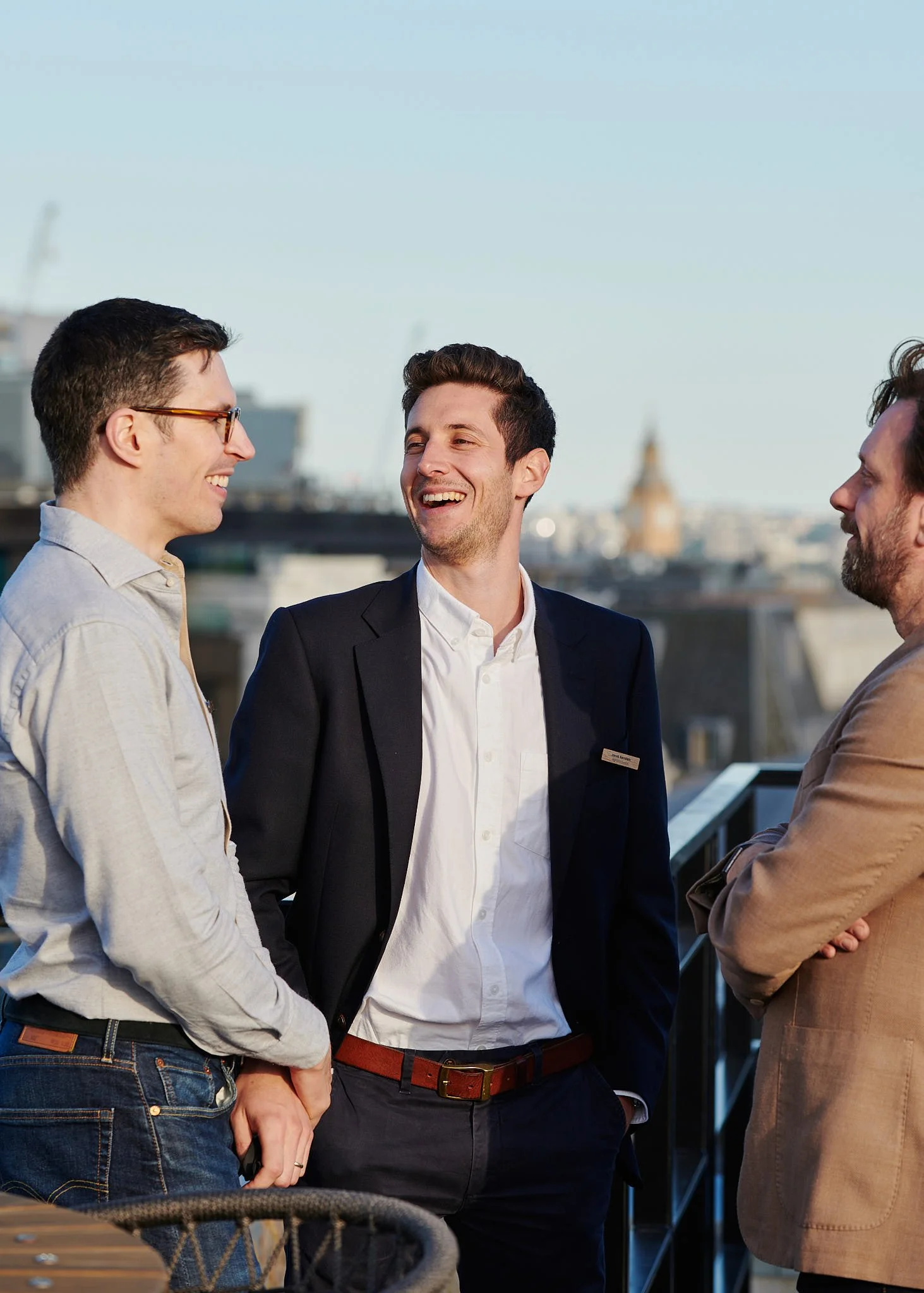 Three men chatting and smiling on a rooftop terrace in a city, with the London skyline in the background.