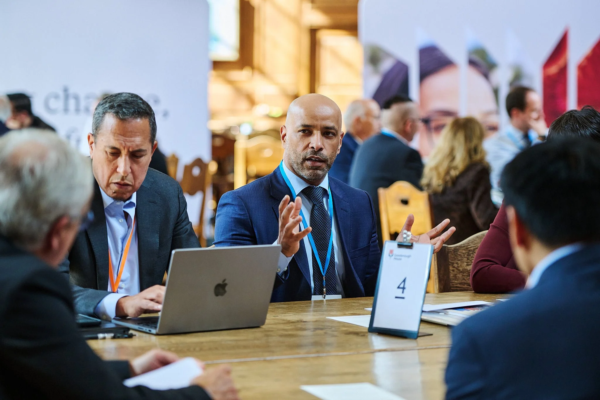 A group of professionals at a conference table, with a man in a blue suit speaking and gesturing, while others listen and work on laptops.