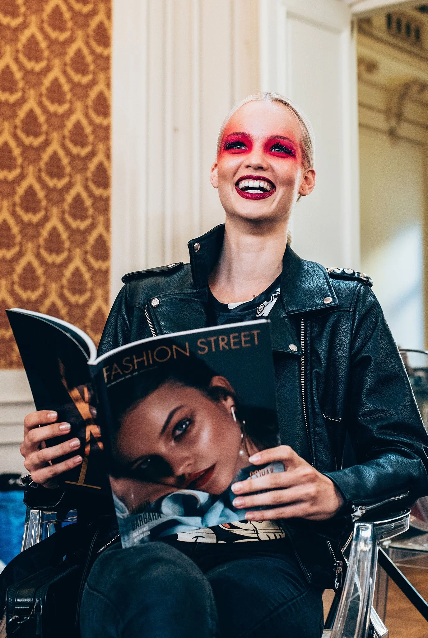 A smiling woman with blonde hair and colorful eye makeup sitting indoors, holding a magazine titled 'Fashion Street' featuring a female model on the cover, wearing a black leather jacket.