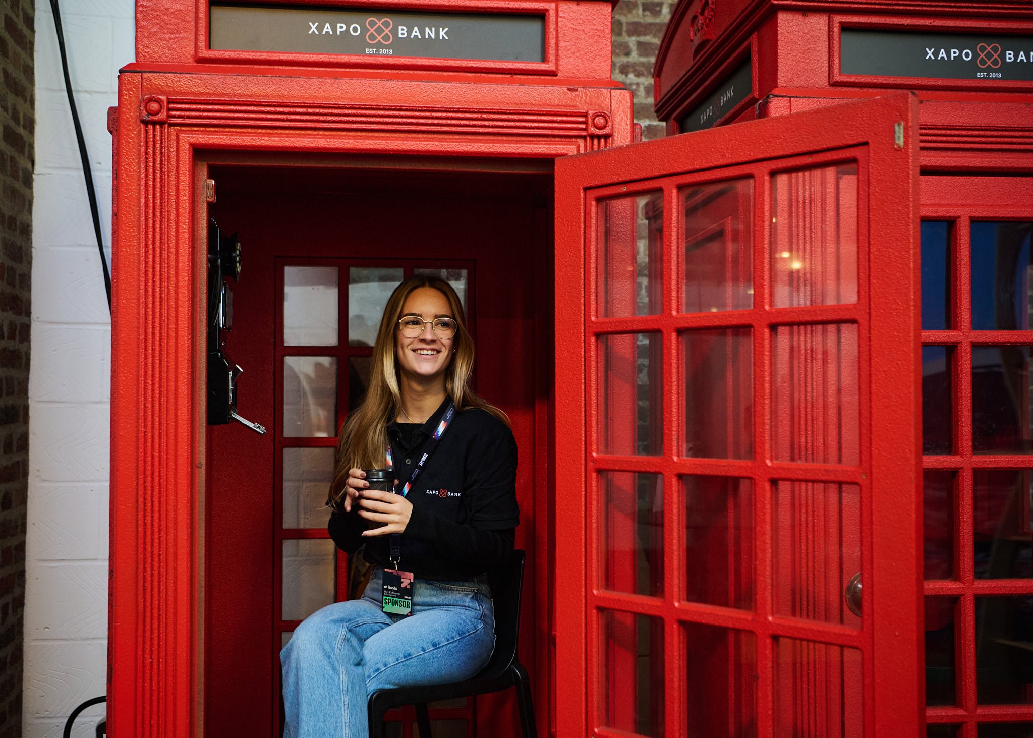 A woman sitting inside a red British telephone booth, smiling, holding a coffee cup, with an ID badge hanging around her neck.