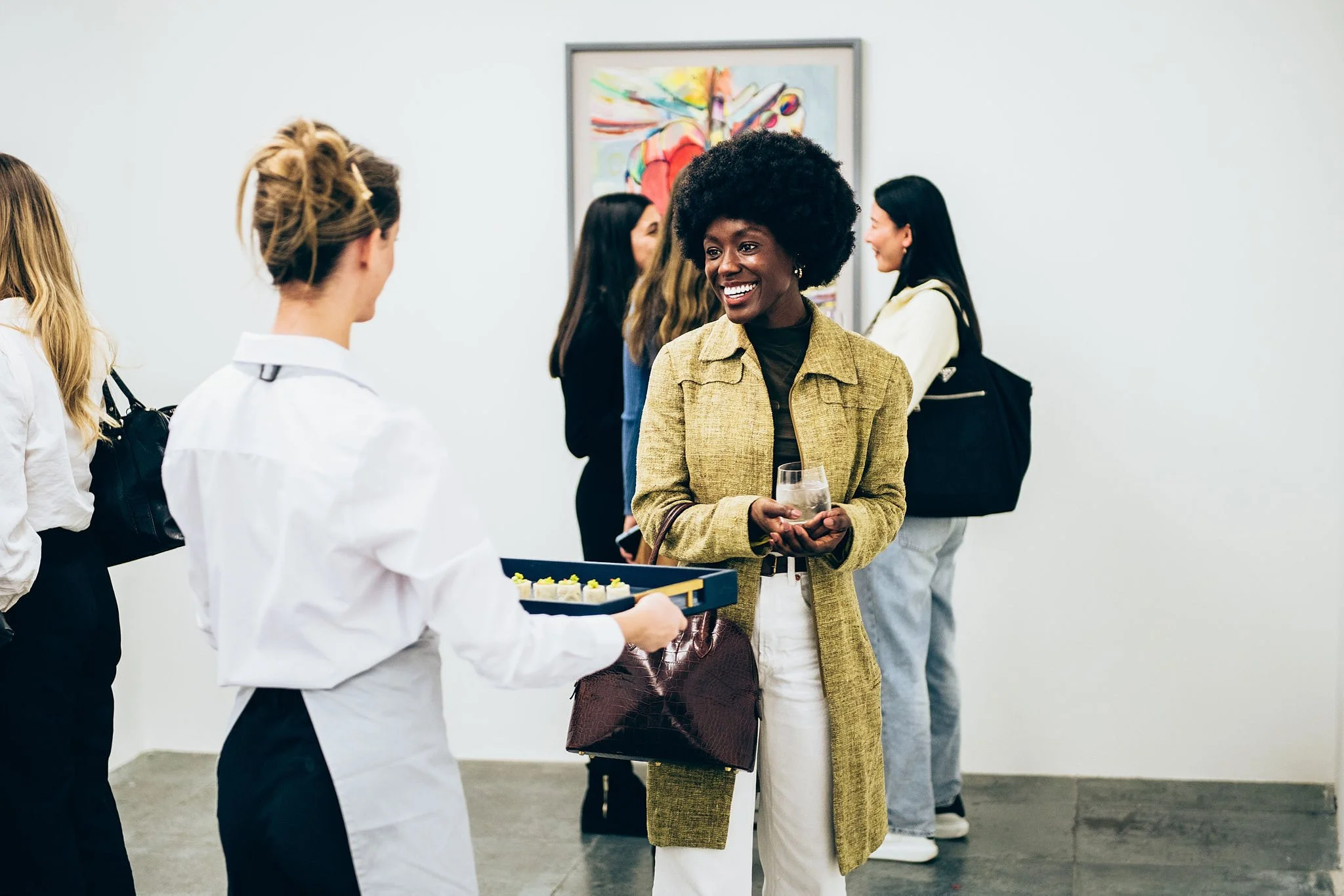 Women chatting at an art gallery, one woman holding a drink, others standing near a painting.