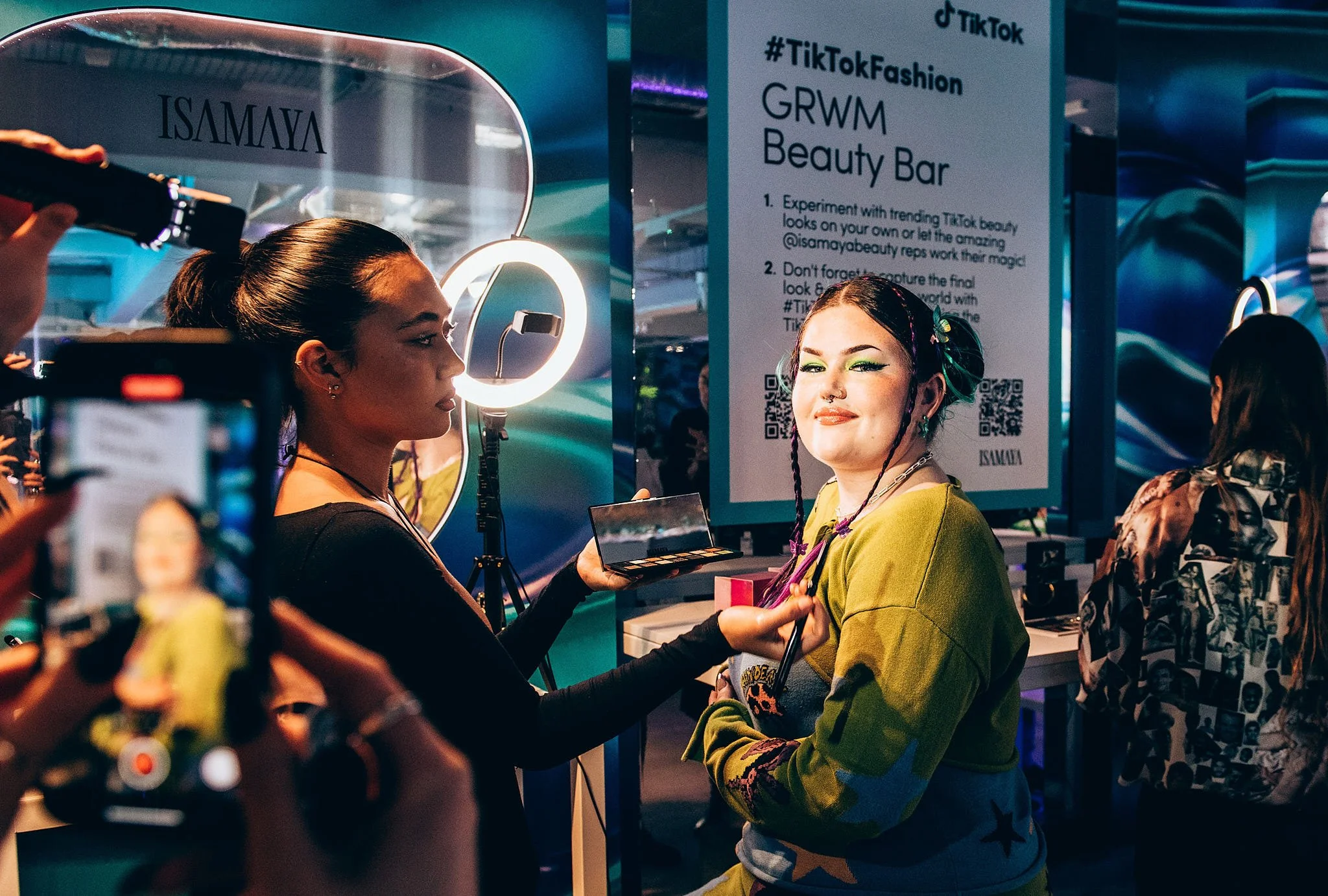 A young woman with colorful makeup and braided hair smiling at a beauty bar station at the TikTok event, with a makeup artist applying eye makeup and another person taking photos.