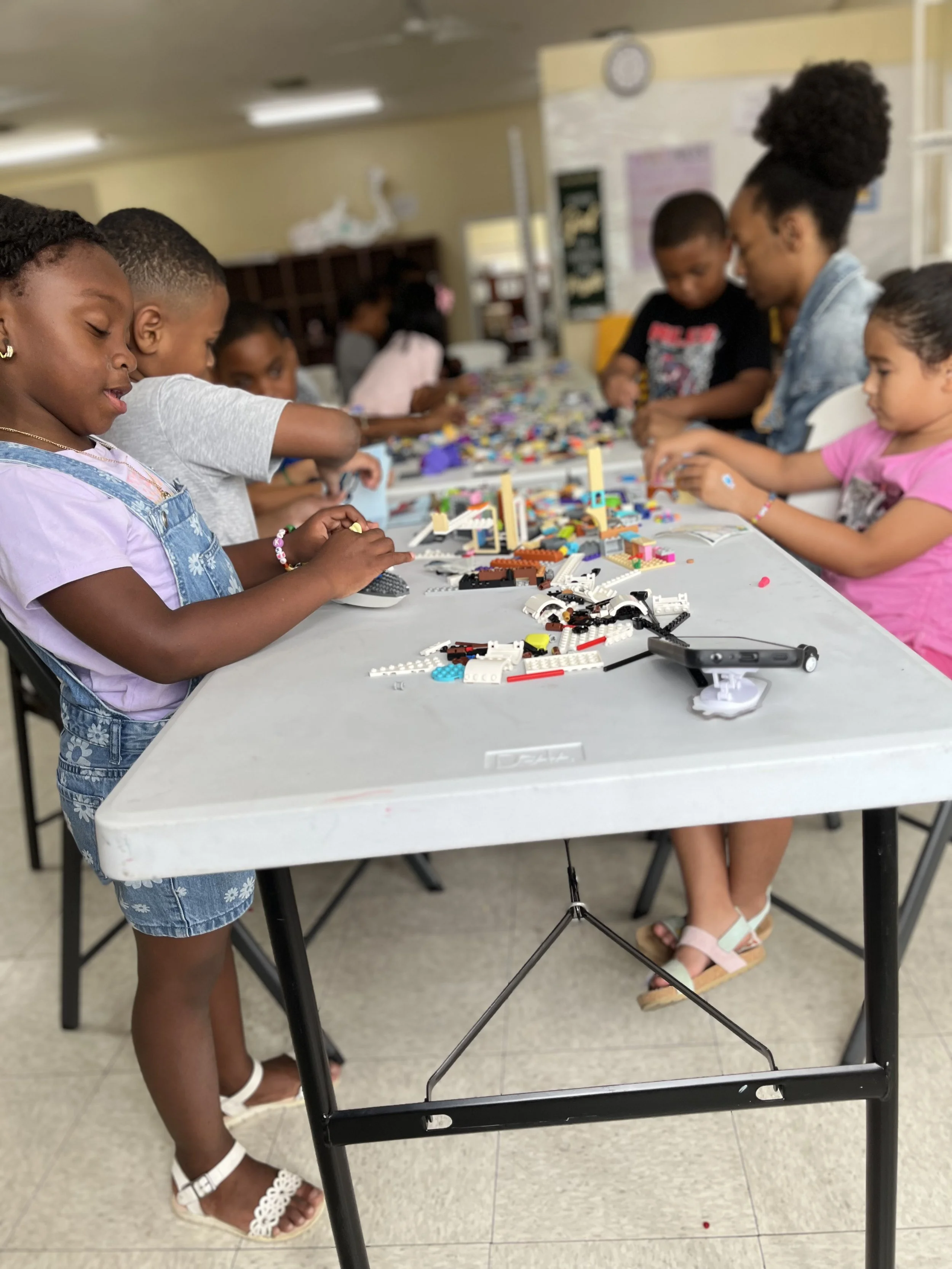 Children and an adult working on Lego building projects at a table in a classroom or community center.