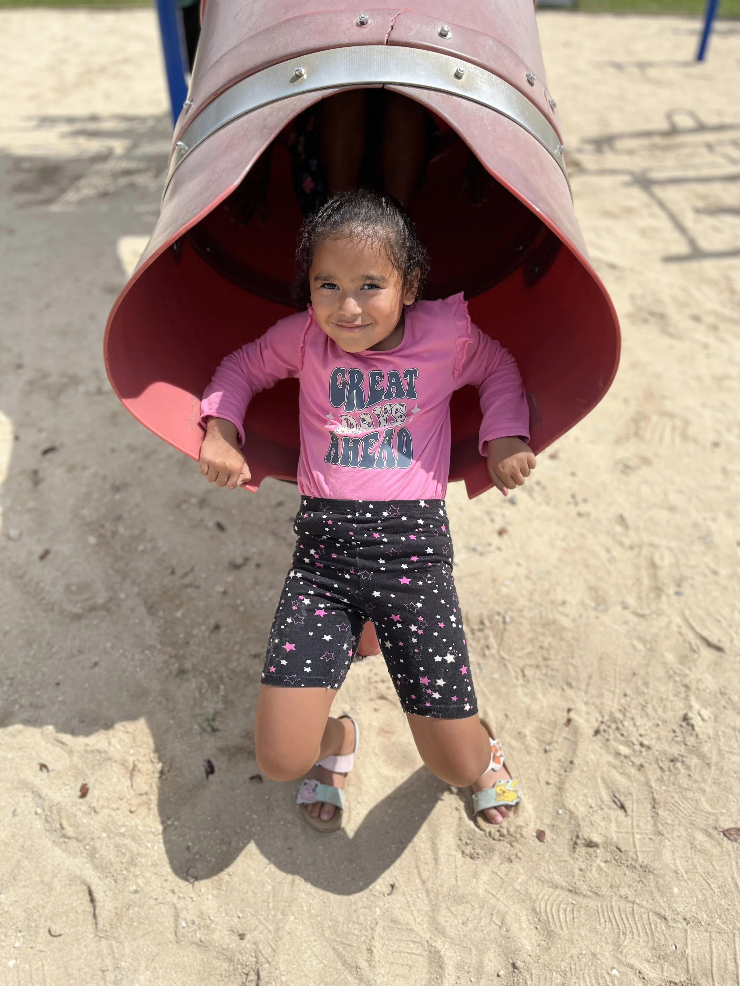 Young girl with dark curly hair and a smiling face, sitting inside a tunnel at a playground, wearing a pink shirt with the words 'Great Days Ahead' and star-patterned shorts, on sandy ground.