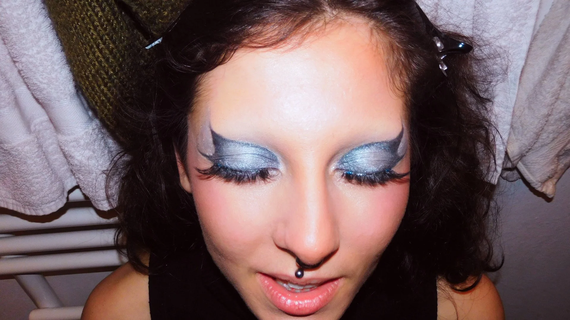 Close-up of a woman with closed eyes, wearing dramatic silver and black eye makeup, long eyelashes, and a septum piercing, with towels in the background.