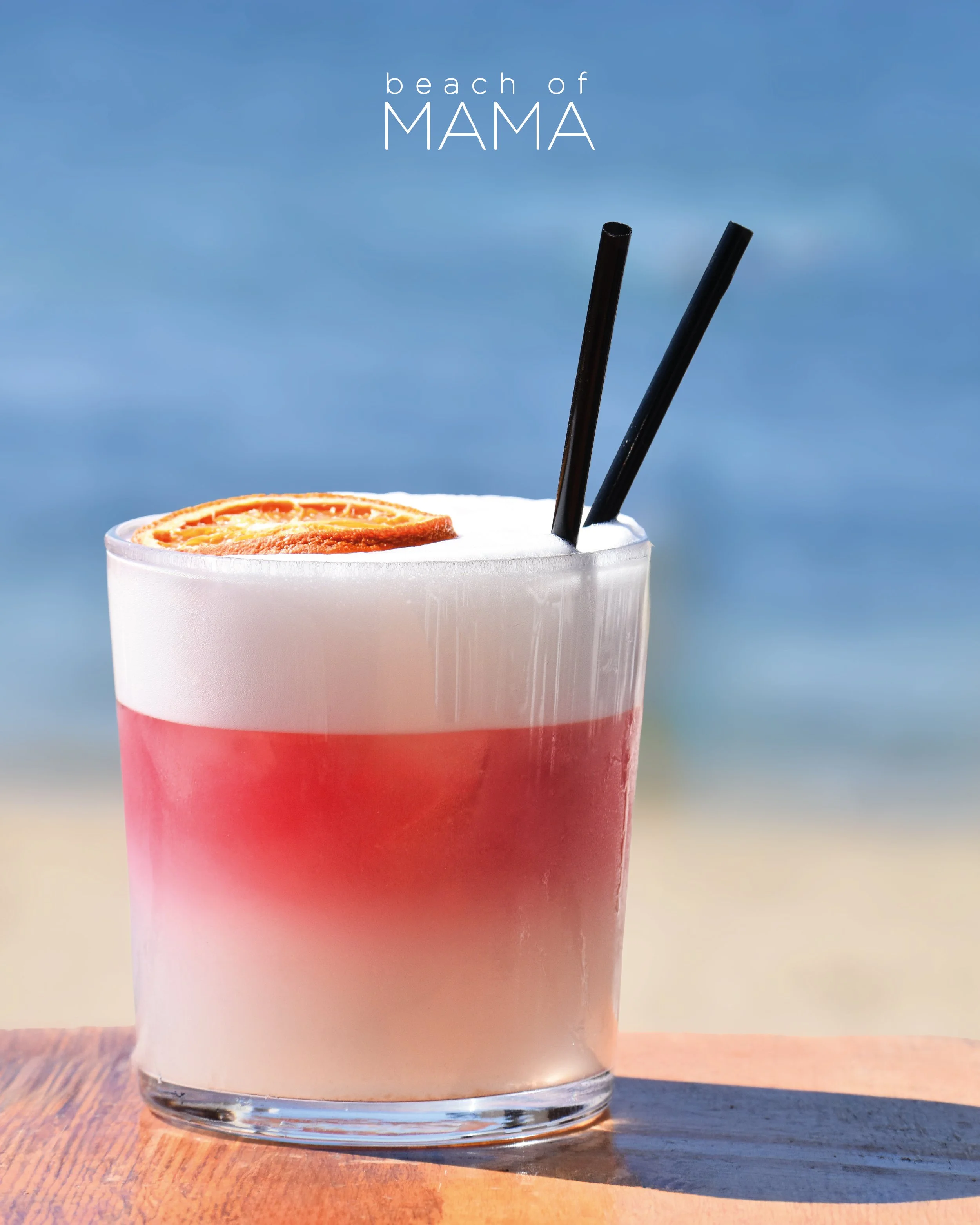 A tropical cocktail with pink and white layers, garnished with a lime wheel, served on a wooden table at the beach, with the ocean in the background.