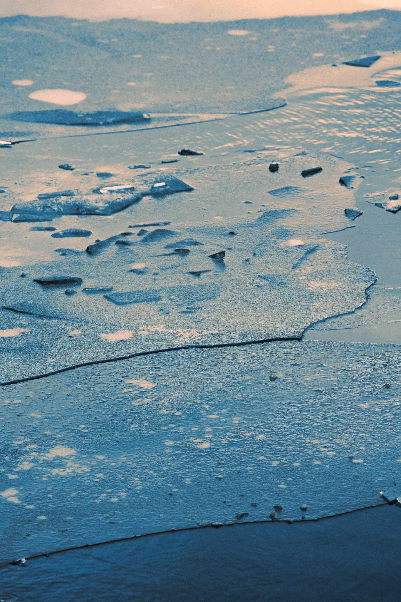 Close-up of a frozen surface with cracks and scattered chunks of ice, with a calm water body partially visible at the bottom.