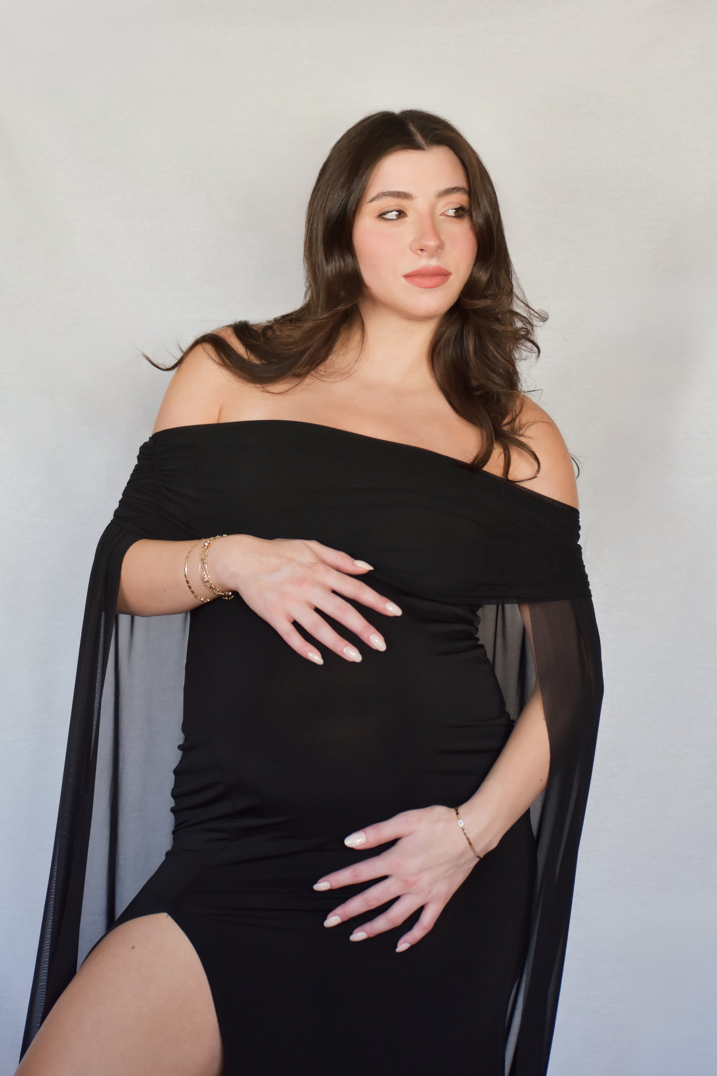 A woman with long brown hair wearing a black off-shoulder dress, posing against a plain background.