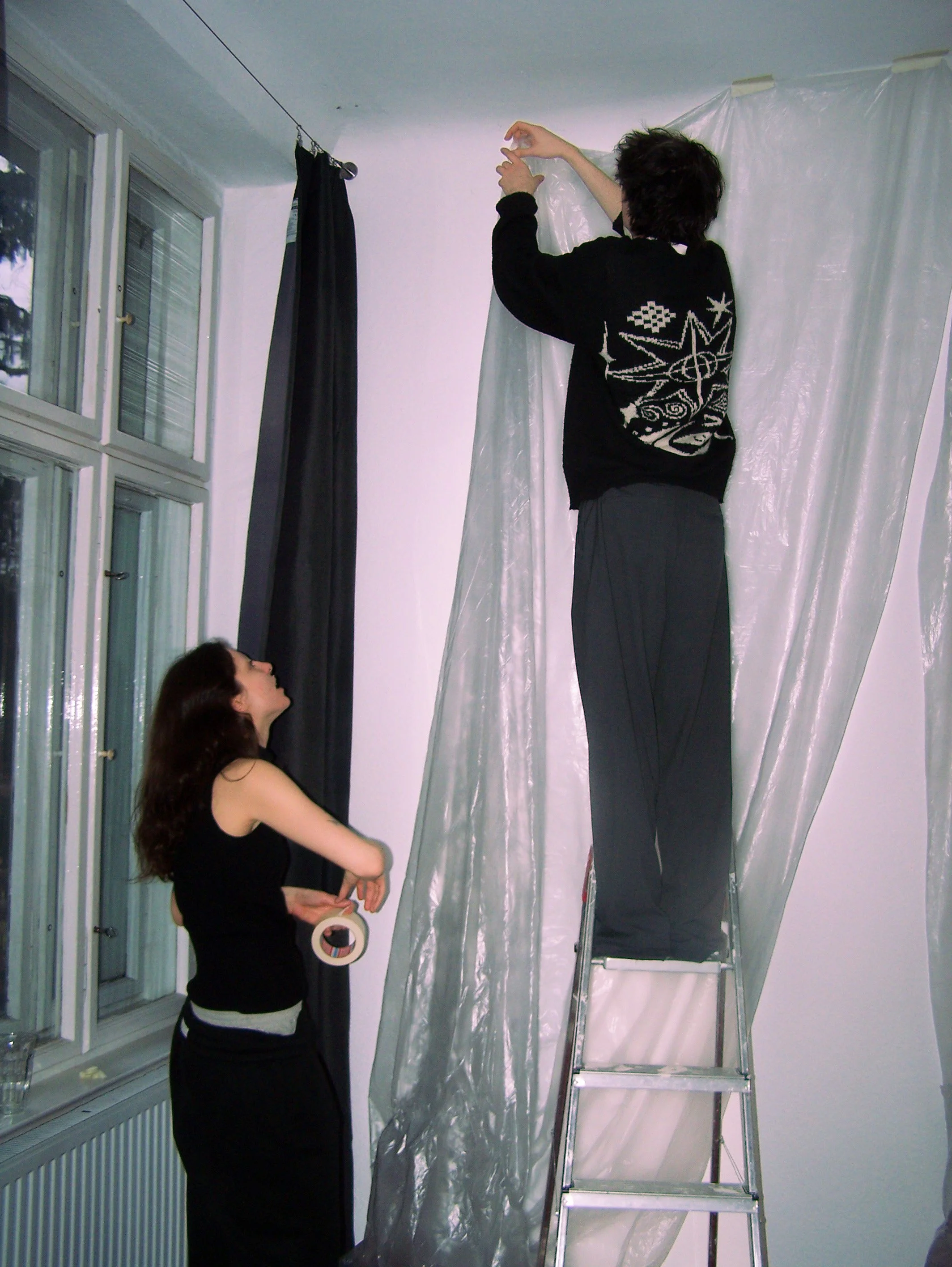 Two people hanging large curtains or fabric panels on a window or wall, one standing on a ladder and the other holding a roll of tape.