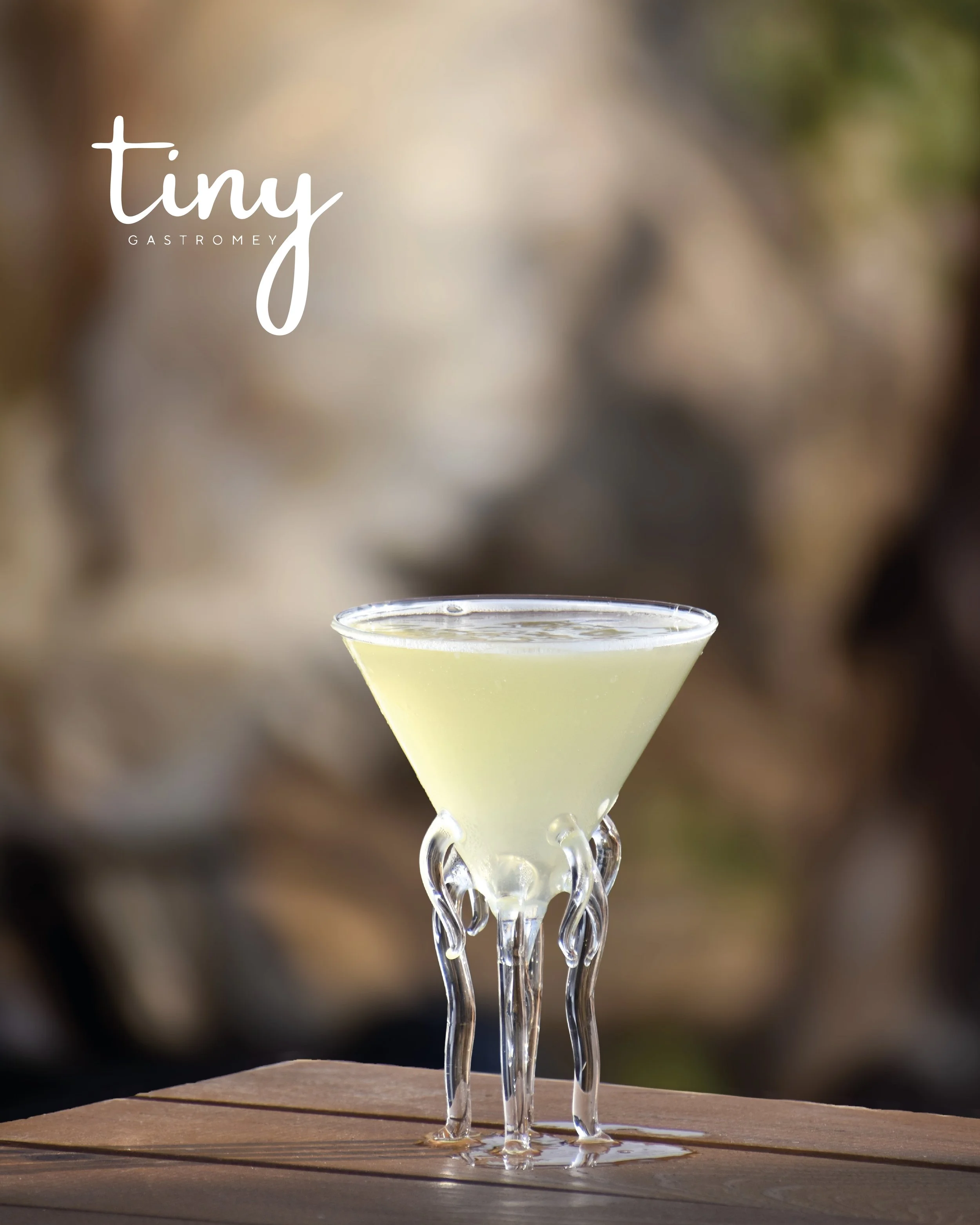 A cocktail in a clear martini glass with a twisted stem, filled with a pale yellow drink, placed on a wooden surface with a blurred outdoor background.