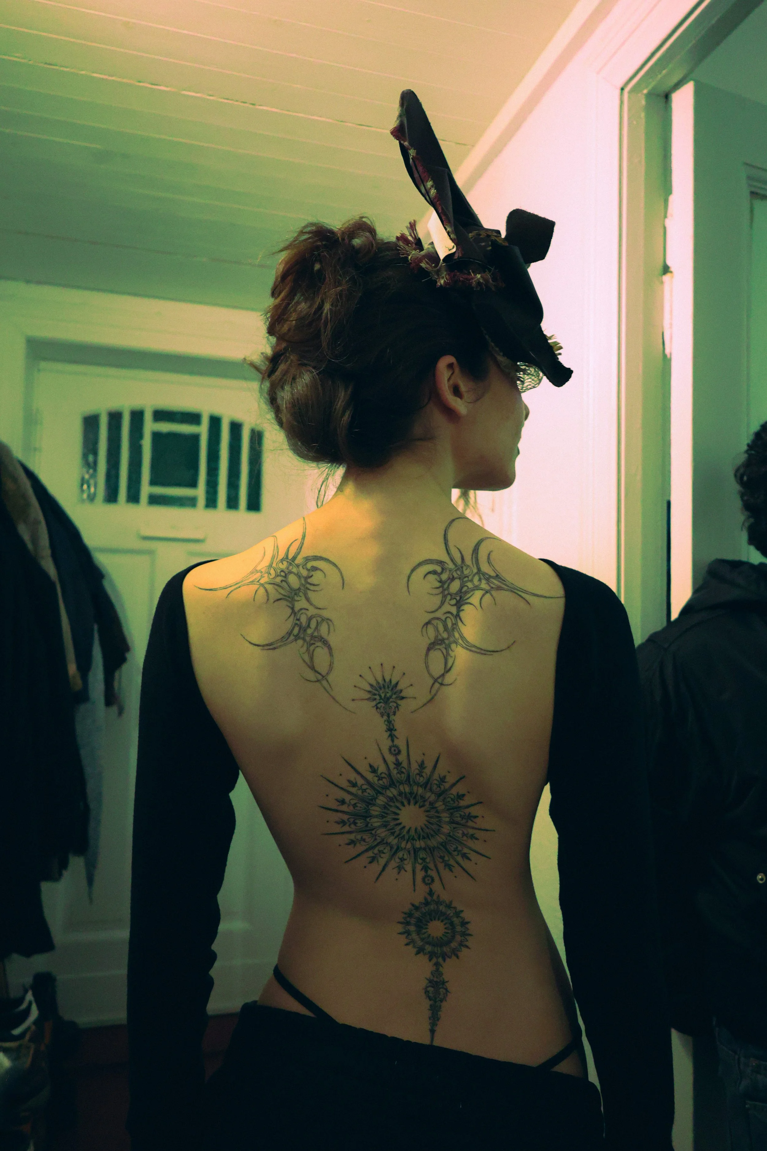 Back of a woman with large black hat, tattooed back with elaborate symmetrical black ink designs.